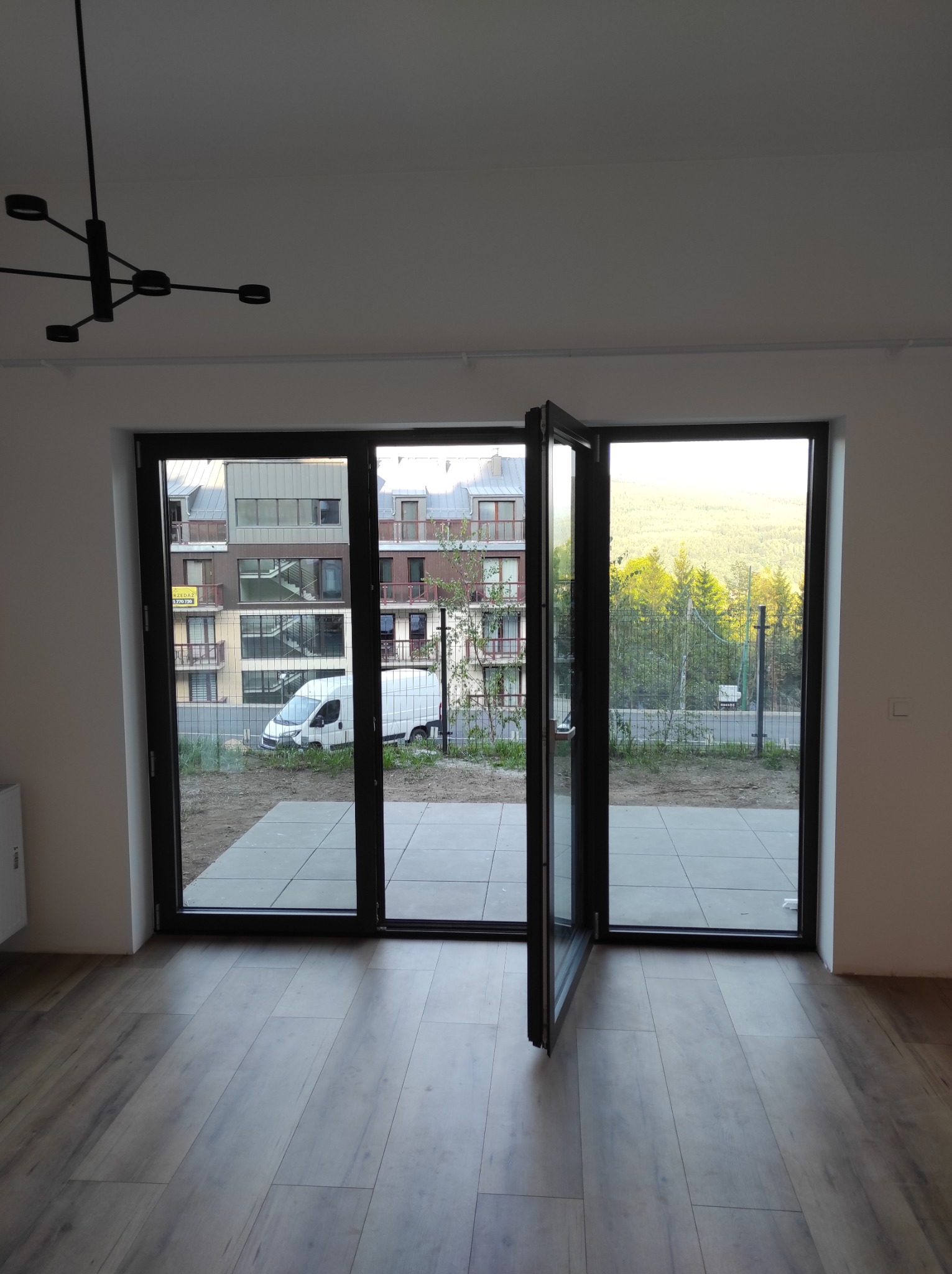 Nowoczesne, trzyskrzydłowe drzwi balkonowe z czarnymi ramami, jedno skrzydło otwarte, widok na nowy budynek i zieleń za oknem, panele podłogowe imitujące drewno, minimalistyczna lampa sufitowa.