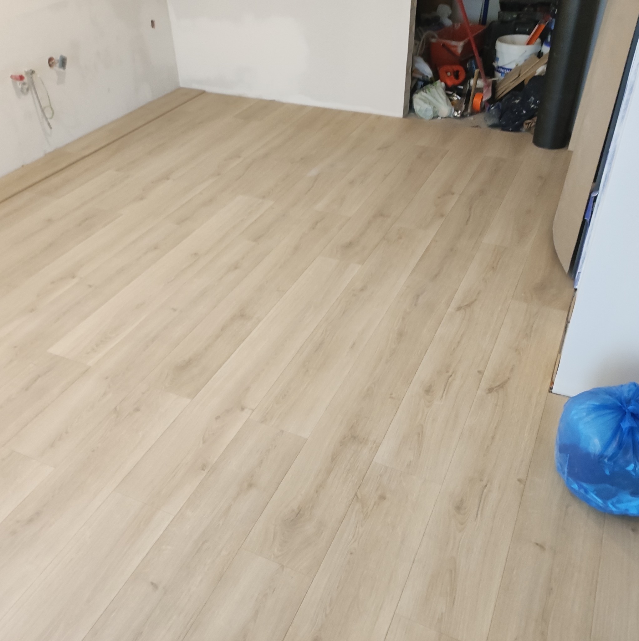 Fragment pokoju z ułożonymi panelami podłogowymi w jasnym odcieniu drewna; widoczna ściana w stanie surowym z wystającymi przewodami i niebieski worek na śmieci w rogu.