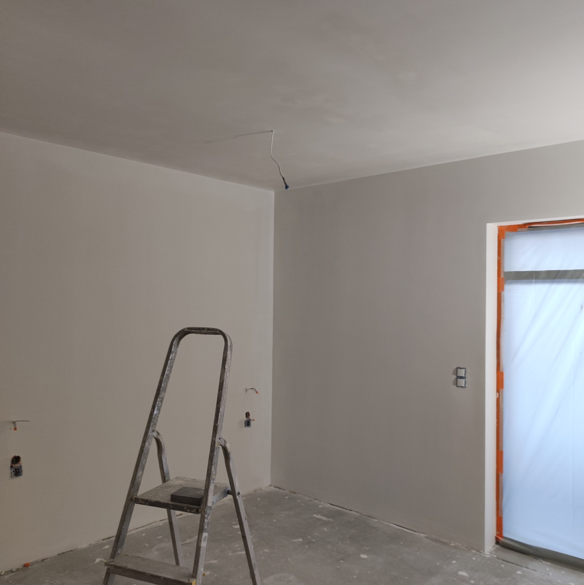 Wnętrze w trakcie remontu: świeżo pomalowane ściany na szaro, aluminiowa drabina, zabezpieczone folią drzwi, widoczne gniazdka elektryczne bez osprzętu.