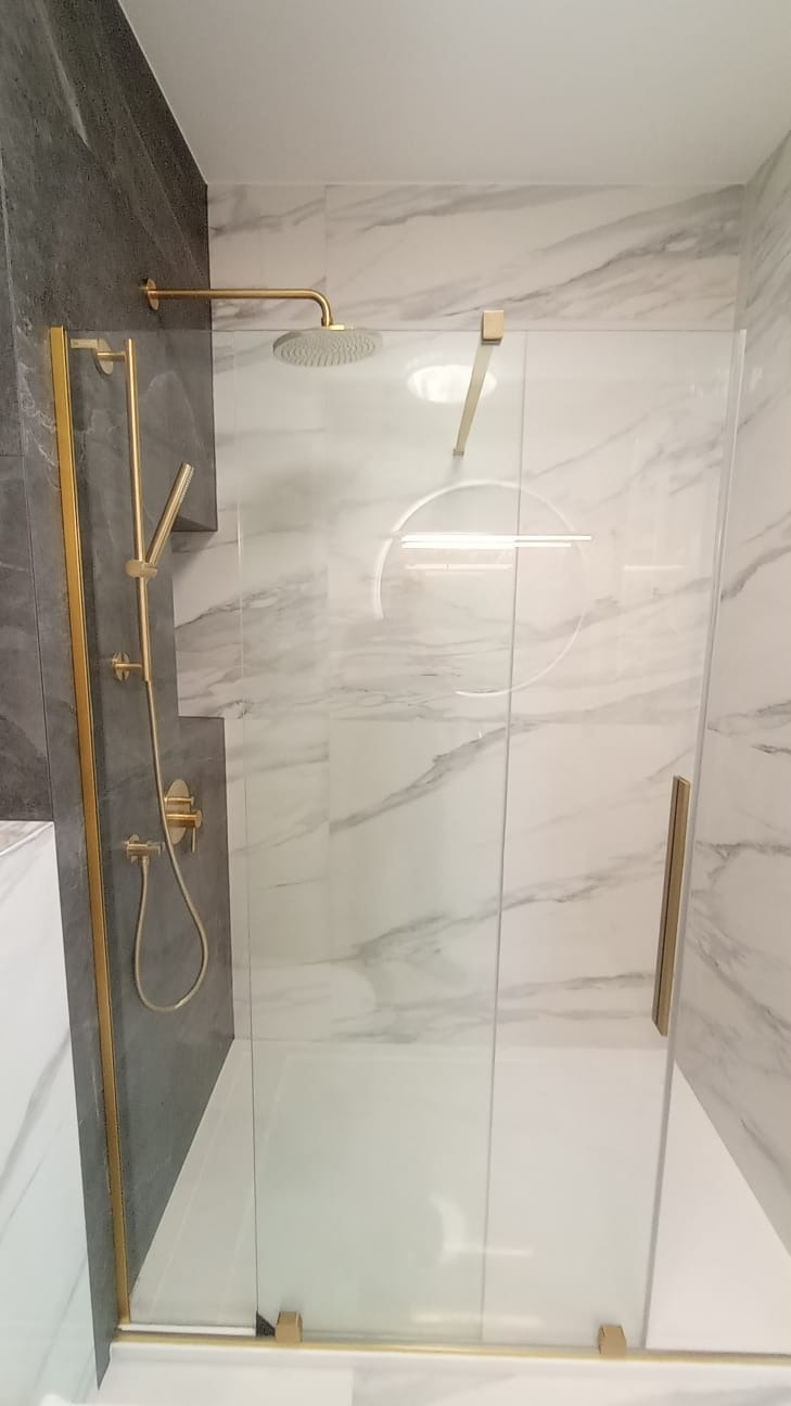 Nowoczesna kabina prysznicowa z złotymi elementami, marmurowe płytki na ścianach, szklane drzwi przesuwne.