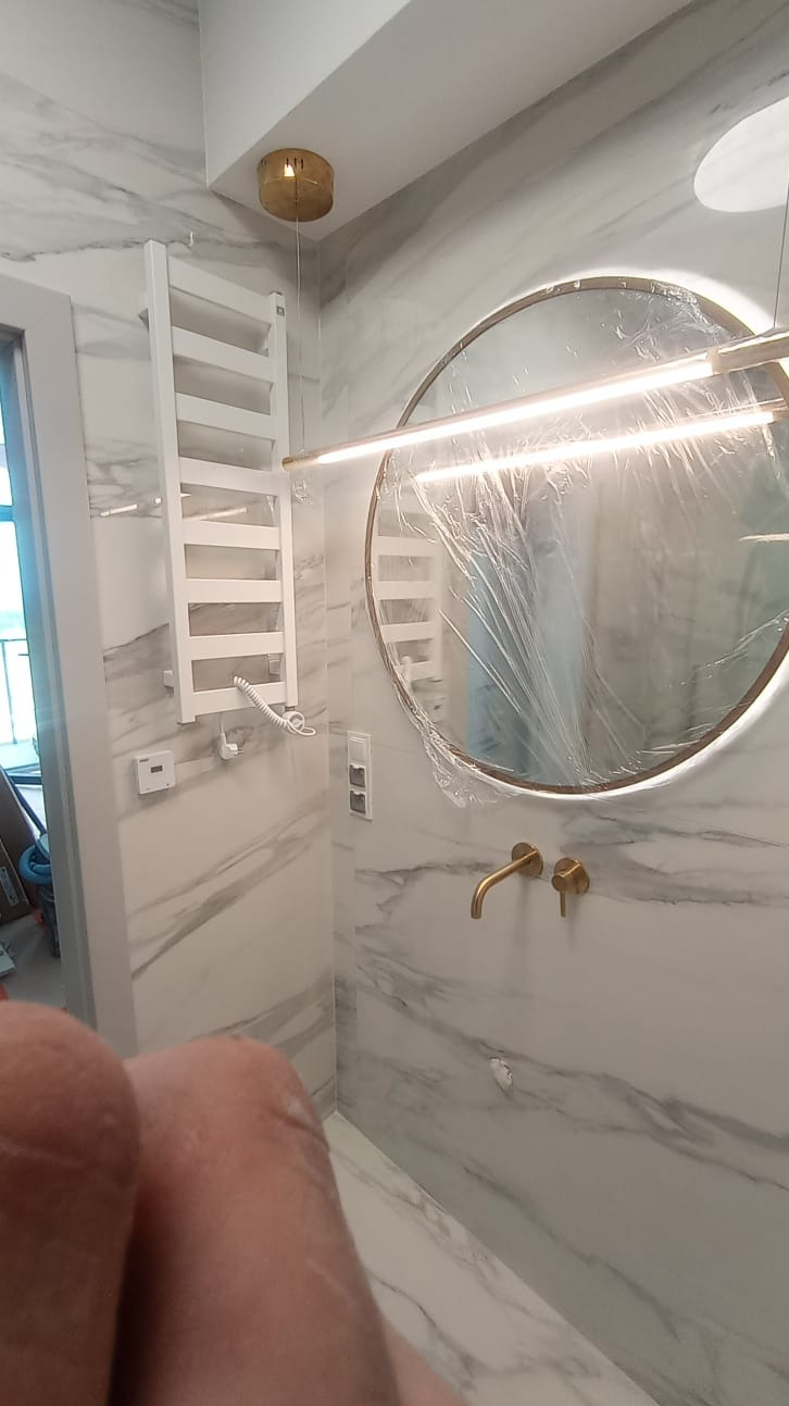 Wykończona łazienka z marmurowymi płytkami, okrągłym lustrem z podświetleniem LED, złotą armaturą i białym grzejnikiem drabinkowym, lustro zabezpieczone folią ochronną.