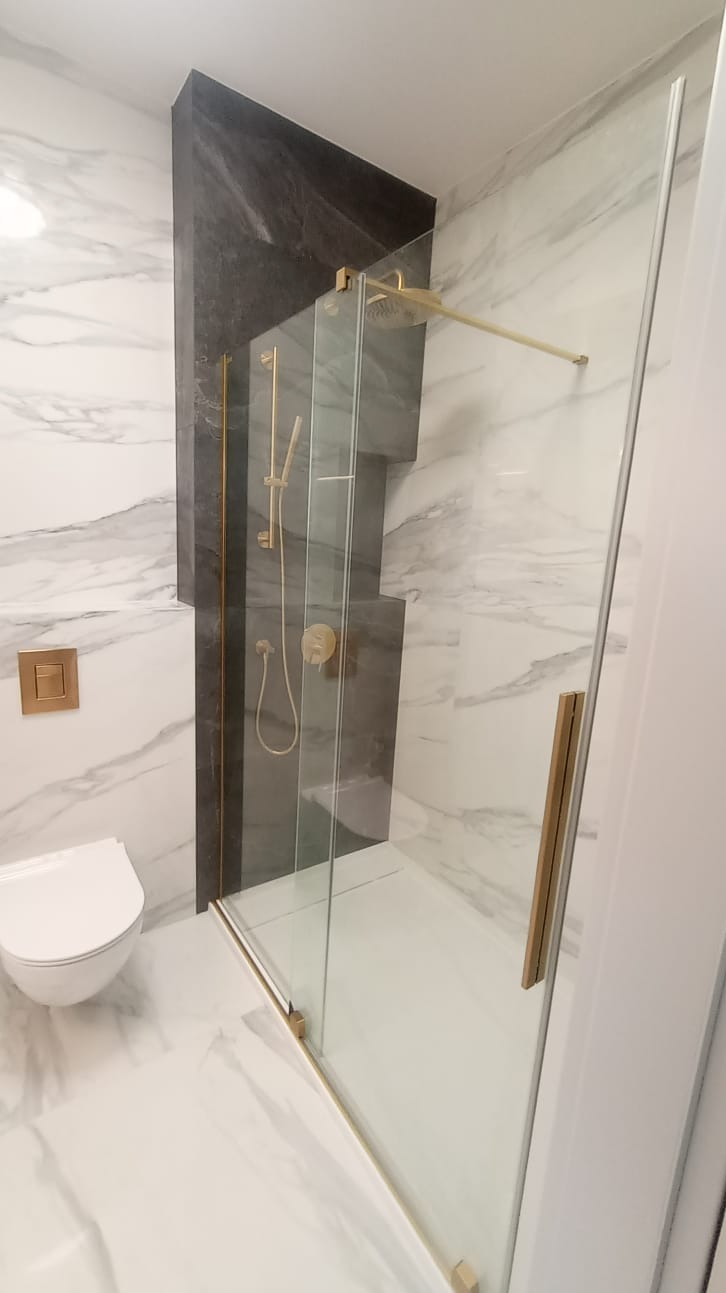 Nowoczesna łazienka z kabiną prysznicową typu walk-in, wykończona marmurowymi płytkami i elementami w kolorze złotym, widoczna toaleta podwieszana i szara wnęka prysznicowa.