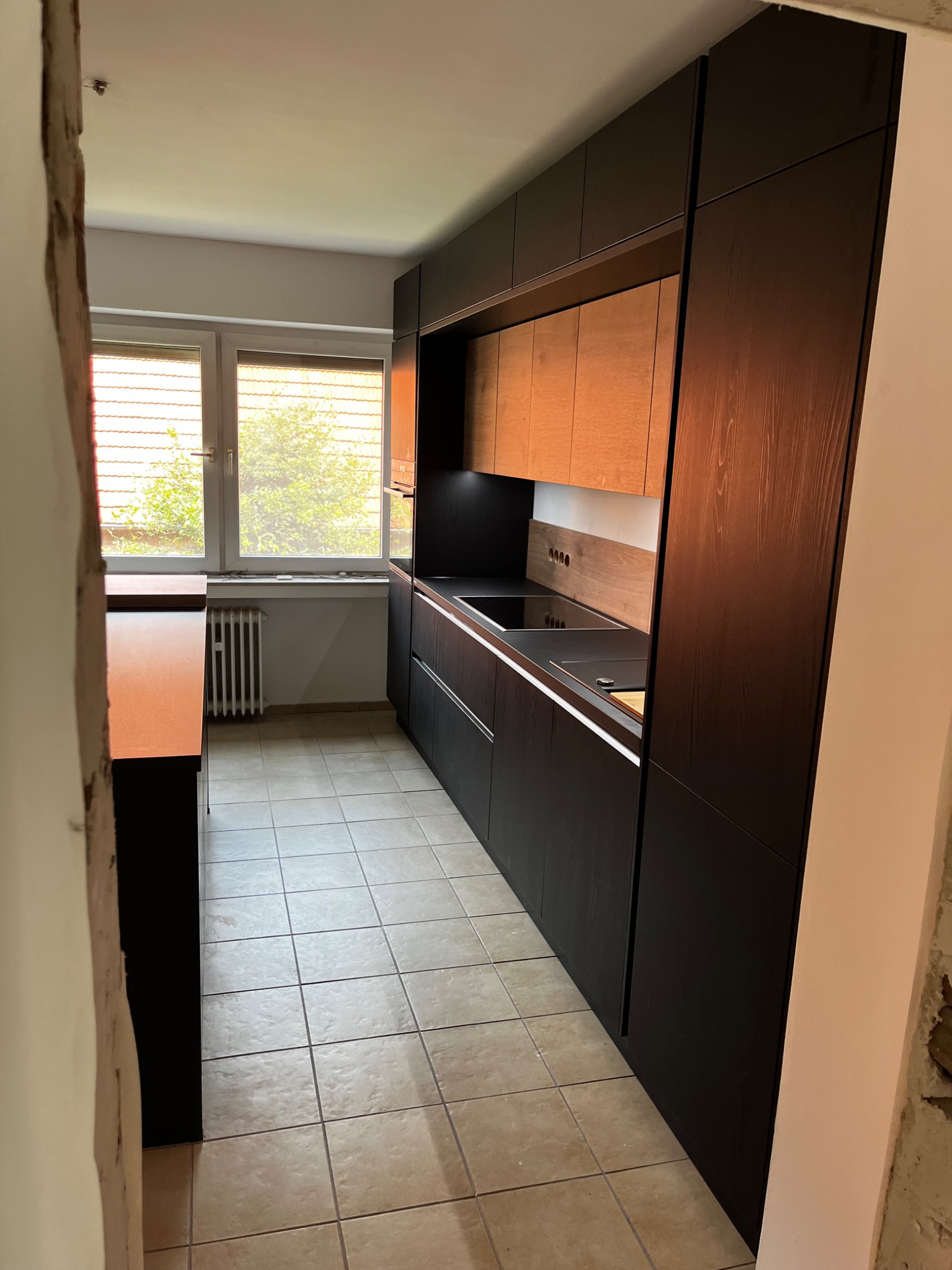 Nowoczesna kuchnia z ciemnymi, drewnianymi szafkami dolnymi i jasnymi, drewnianymi szafkami górnymi, widziana z perspektywy wejścia do pomieszczenia, z widocznym fragmentem niezakończonego...