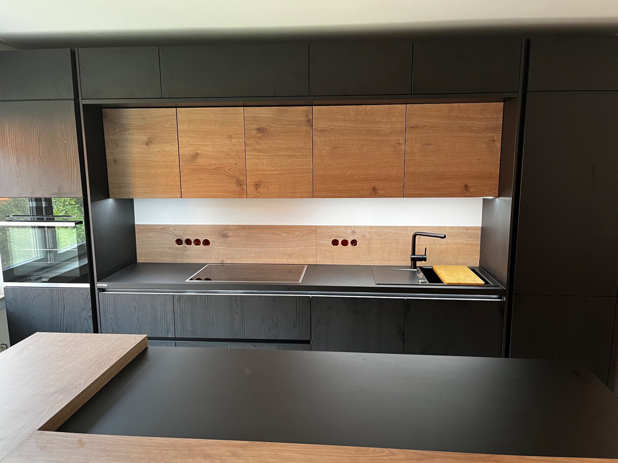 Nowoczesna kuchnia z czarnymi szafkami i drewnianymi frontami górnych szafek, widoczny blat z wbudowaną płytą indukcyjną i zlewem z czarną armaturą oraz drewnianą deską do krojenia.