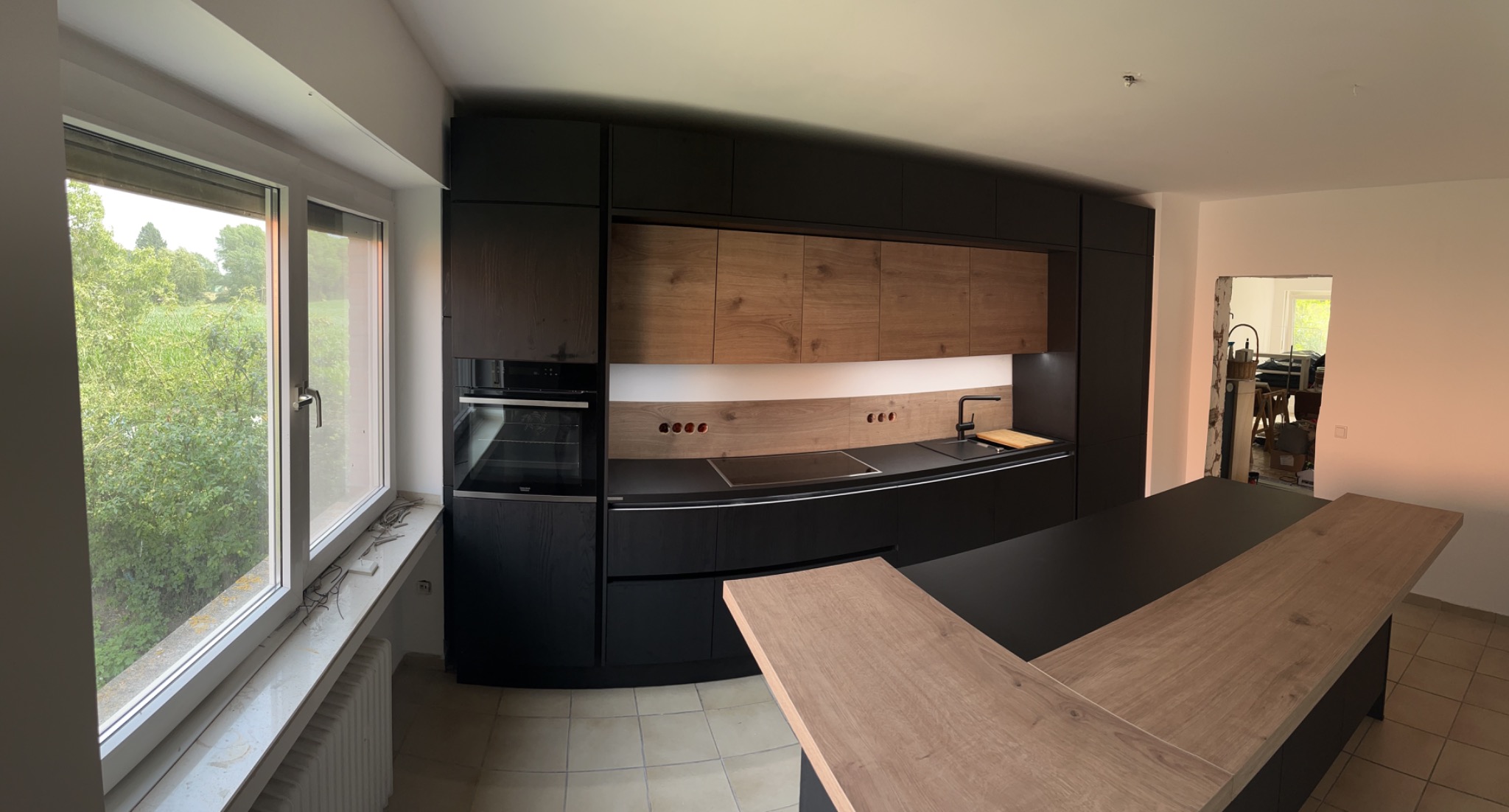 Nowoczesna kuchnia z czarnymi szafkami i drewnianymi akcentami, wyspa z dwoma blatami, widok na ogród przez okno, widoczne gniazdka elektryczne w ścianie.