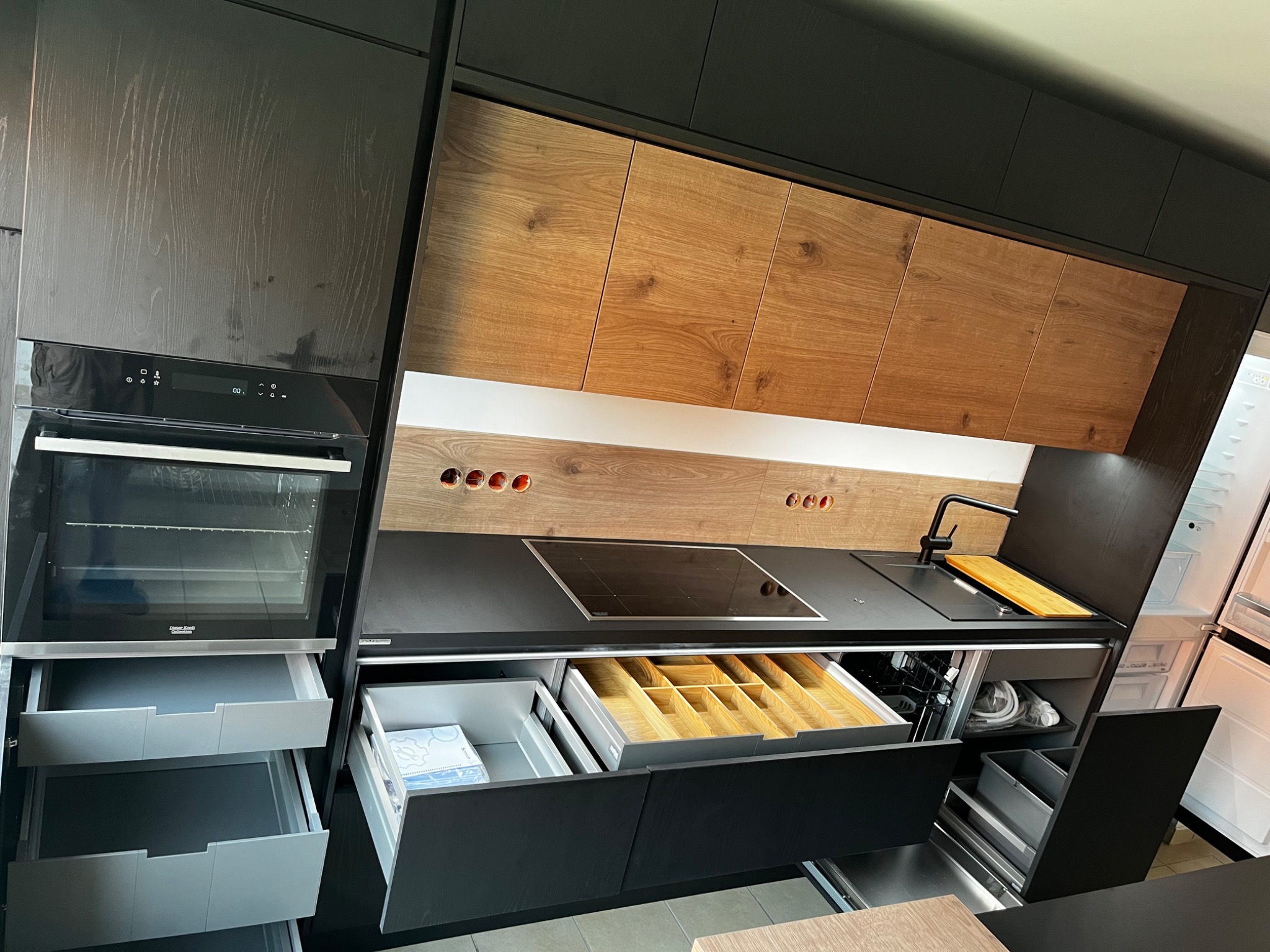 Nowoczesna kuchnia z czarnymi frontami i drewnianymi szafkami górnymi, otwarte szuflady z organizerami na sztućce i akcesoria, widoczny piekarnik i płyta indukcyjna.