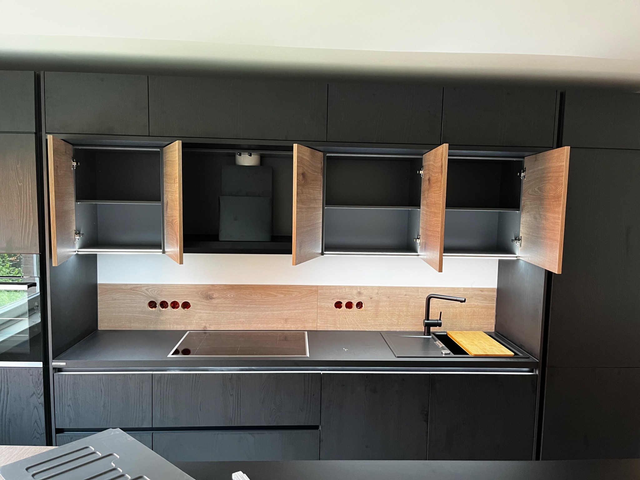 Nowoczesna, czarna zabudowa kuchenna z drewnianymi akcentami, otwarte szafki górne, płyta indukcyjna i czarny kran, widoczne gniazdka elektryczne w panelu nad blatem.