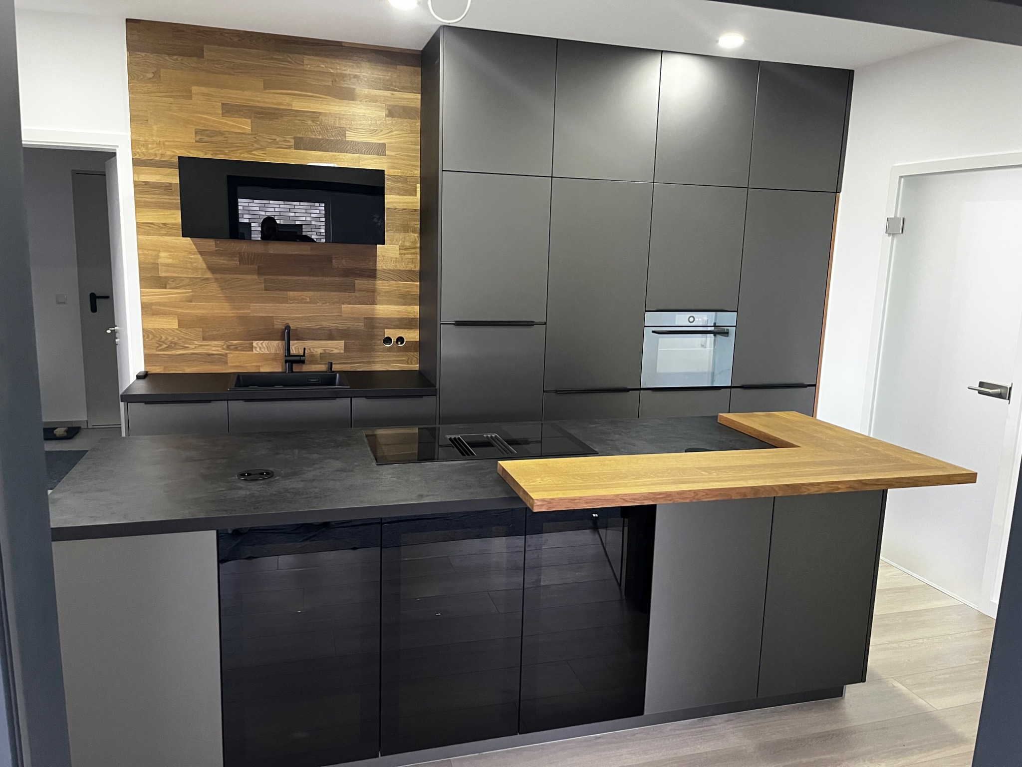 Nowoczesna kuchnia z szarymi, matowymi szafkami, drewnianą ścianą i wyspą z blatem z ciemnego kamienia i drewnianym przedłużeniem.
