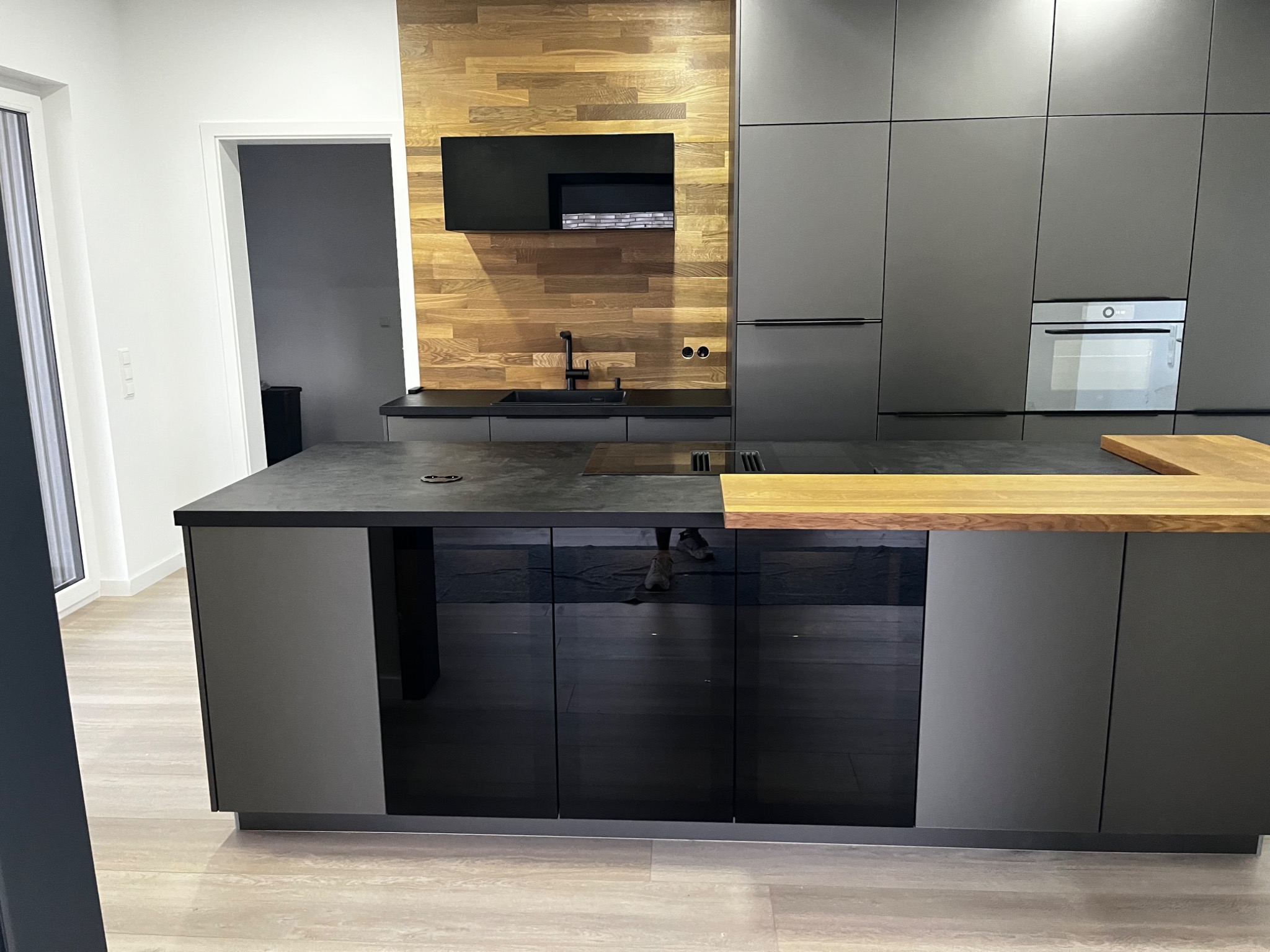 Nowoczesna kuchnia z matowymi szarymi szafkami, drewnianym panelem ściennym i czarnym sprzętem, wyspa z czarnymi, błyszczącymi frontami i blatem z drewna.