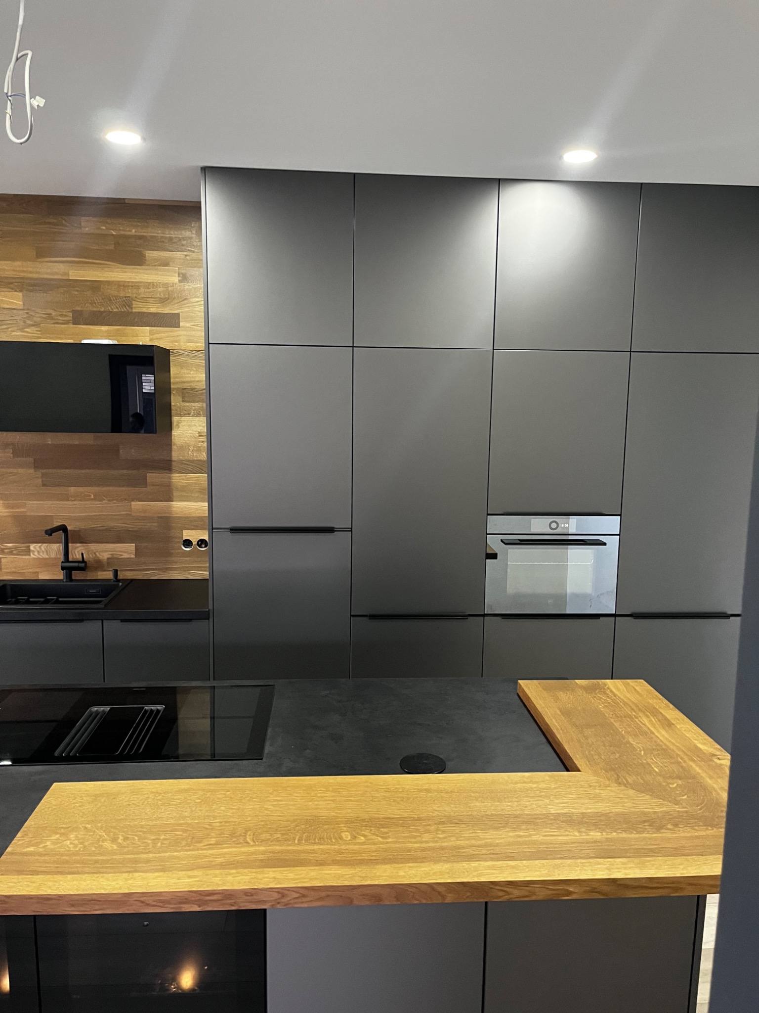 Nowoczesna kuchnia w odcieniach szarości z drewnianym blatem wyspowym i elementami z naturalnego drewna na ścianie, minimalistyczny design z wbudowanym sprzętem AGD.