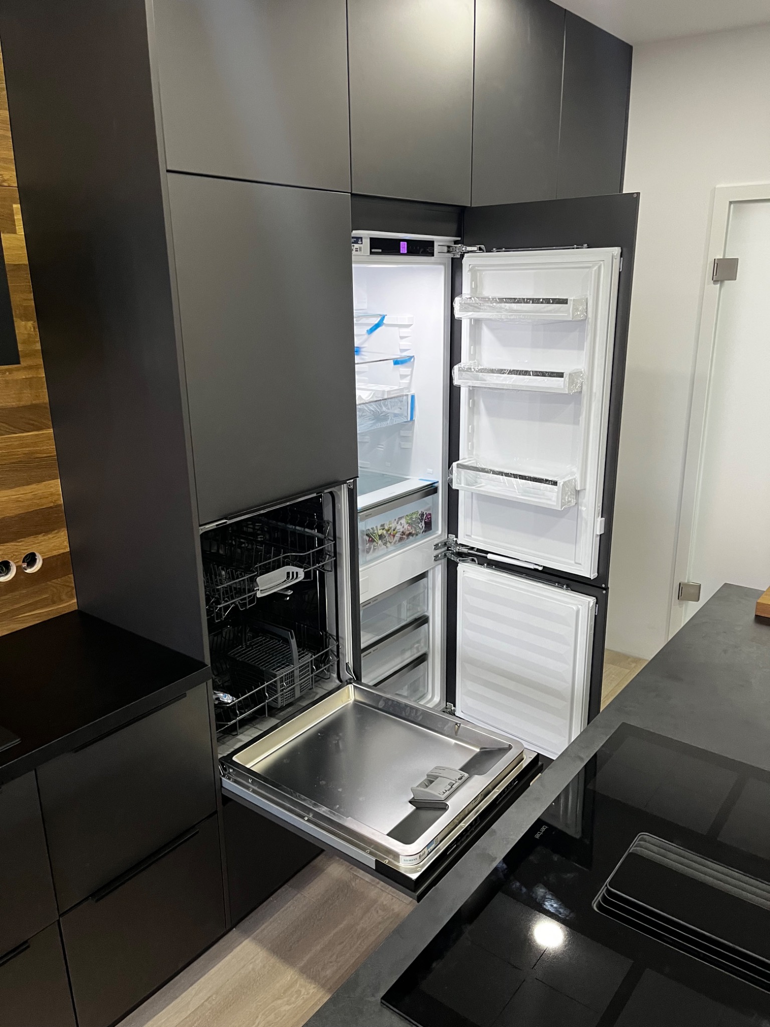 Czarna, nowoczesna zabudowa kuchenna z otwartą lodówką i zmywarką z wysuniętą szufladą na naczynia. Widoczne drewniane panele na ścianie i płyta indukcyjna na blacie.