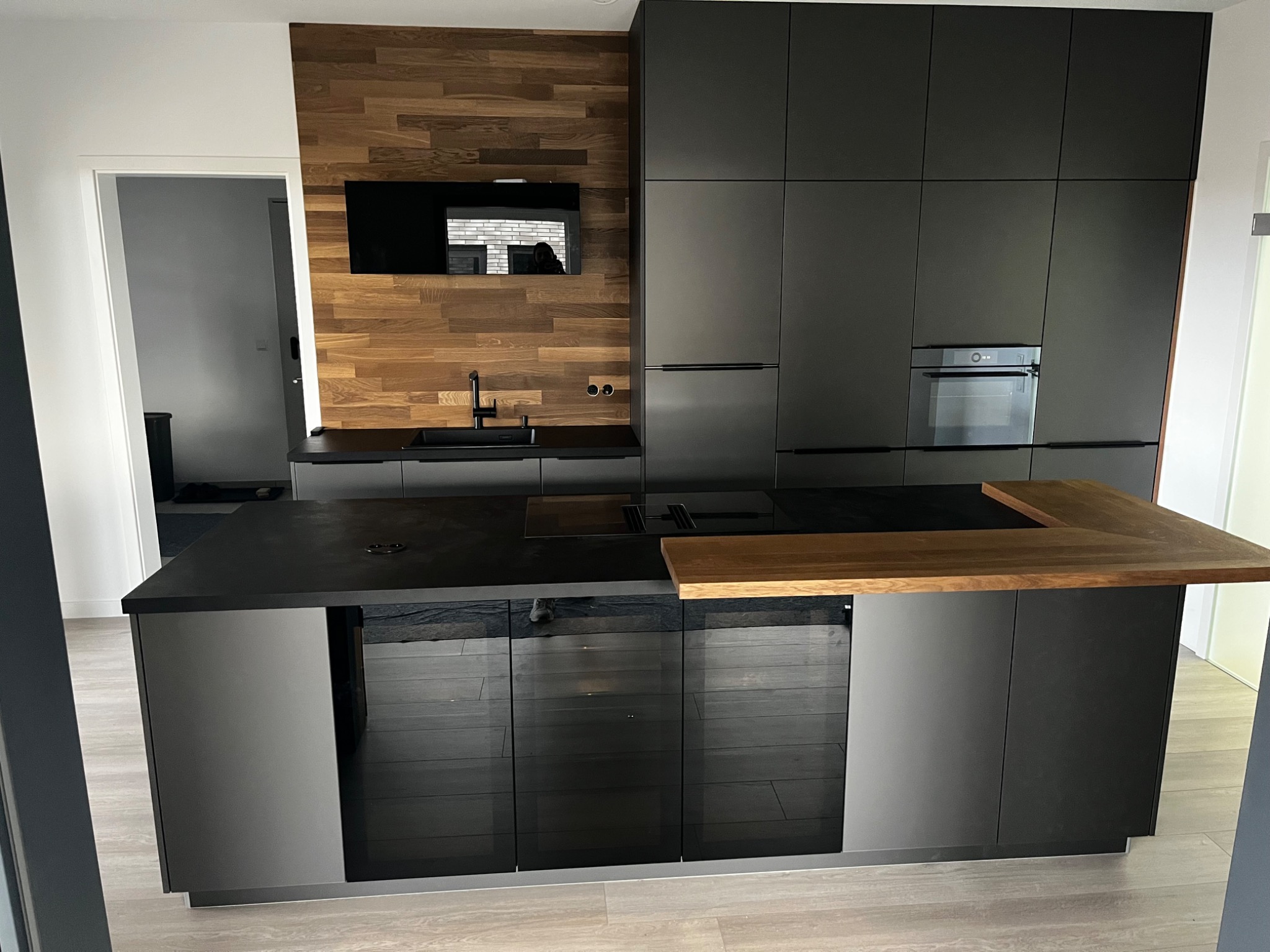 Nowoczesna kuchnia z czarnymi szafkami i drewnianym panelem ściennym, wyspa z drewnianym blatem i przeszklonymi frontami szafek.