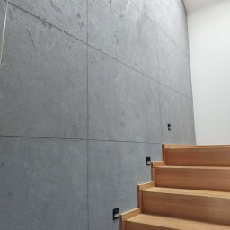 Termmodern - Ściana z szarego betonu architektonicznego ułożonego w kwadraty, oświetlenie schodowe przy drewnianych schodach, naturalne światło z góry.