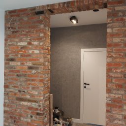 Termmodern - Aranżacja wnętrza z ceglanym łukiem nad drzwiami, surowa cegła kontrastuje z gładką, szarą ścianą i białą stolarką drzwiową, widoczne narzędzia i materiały budowlane sugerują etap wykończeniowy.