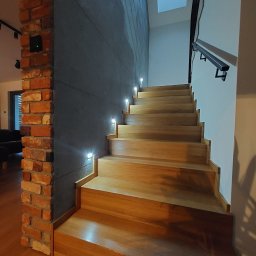 Termmodern - Nowoczesne schody drewniane z oświetleniem punktowym w ścianie o surowym wykończeniu, kolumna z cegły rozbiórkowej po lewej stronie, widok z dołu.