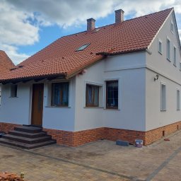 Termmodern - Odnowiony dom jednorodzinny z nową elewacją w kolorze jasnego beżu, czerwoną dachówką i ceglanym cokołem, widok z chodnika z kostki brukowej w słoneczny dzień.