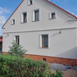 Termmodern - Wykończona elewacja domu z jasnoszarym tynkiem, dekoracyjnymi elementami pod dachem i ceglaną podmurówką, widok z poziomu ogrodu z krzewami i małą choinką.