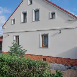 Wykończona elewacja domu z jasnoszarym tynkiem, dekoracyjnymi elementami pod dachem i ceglaną podmurówką, widok z poziomu ogrodu z krzewami i małą choinką.