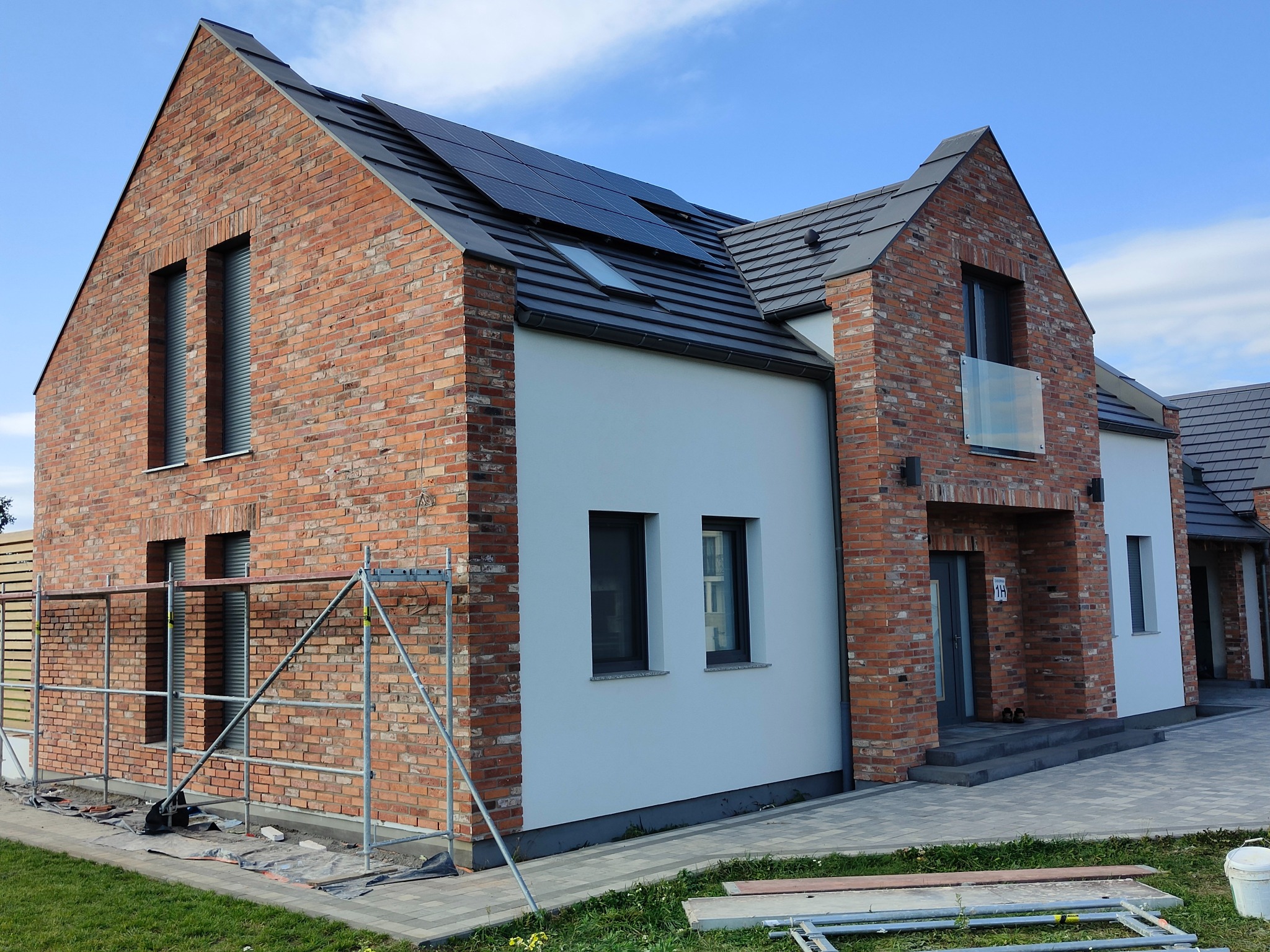 Nowoczesny dom jednorodzinny z elewacją częściowo wykończoną czerwoną cegłą klinkierową, białą elewacją, ciemną dachówką i panelami słonecznymi na dachu, widoczne rusztowanie przy ścianie bocznej.