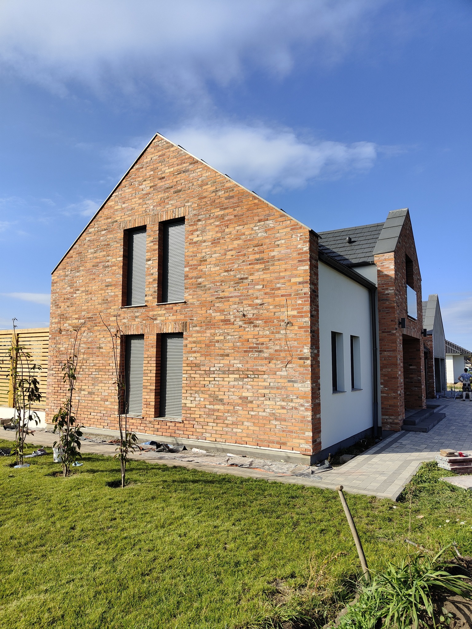 Nowoczesny dom jednorodzinny z elewacją z cegły klinkierowej i jasnymi elementami, widok z trawnika z młodymi drzewkami.