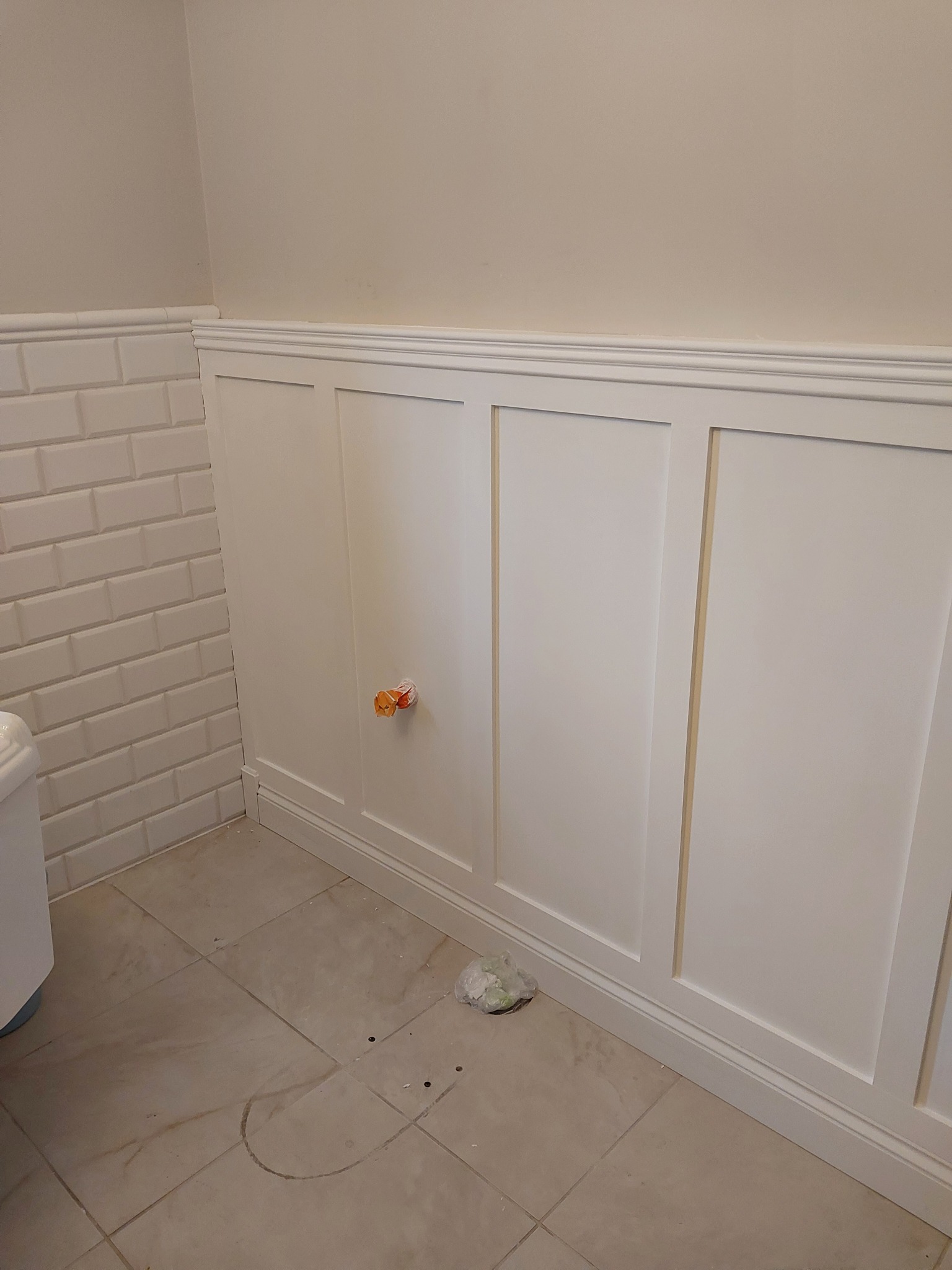 Białe panele ścienne w łazience, częściowo wykończone, widoczne ślady po pracach na podłodze, rura wystająca ze ściany zabezpieczona folią, dolna część ściany wyłożona białymi płytkami typu metro.