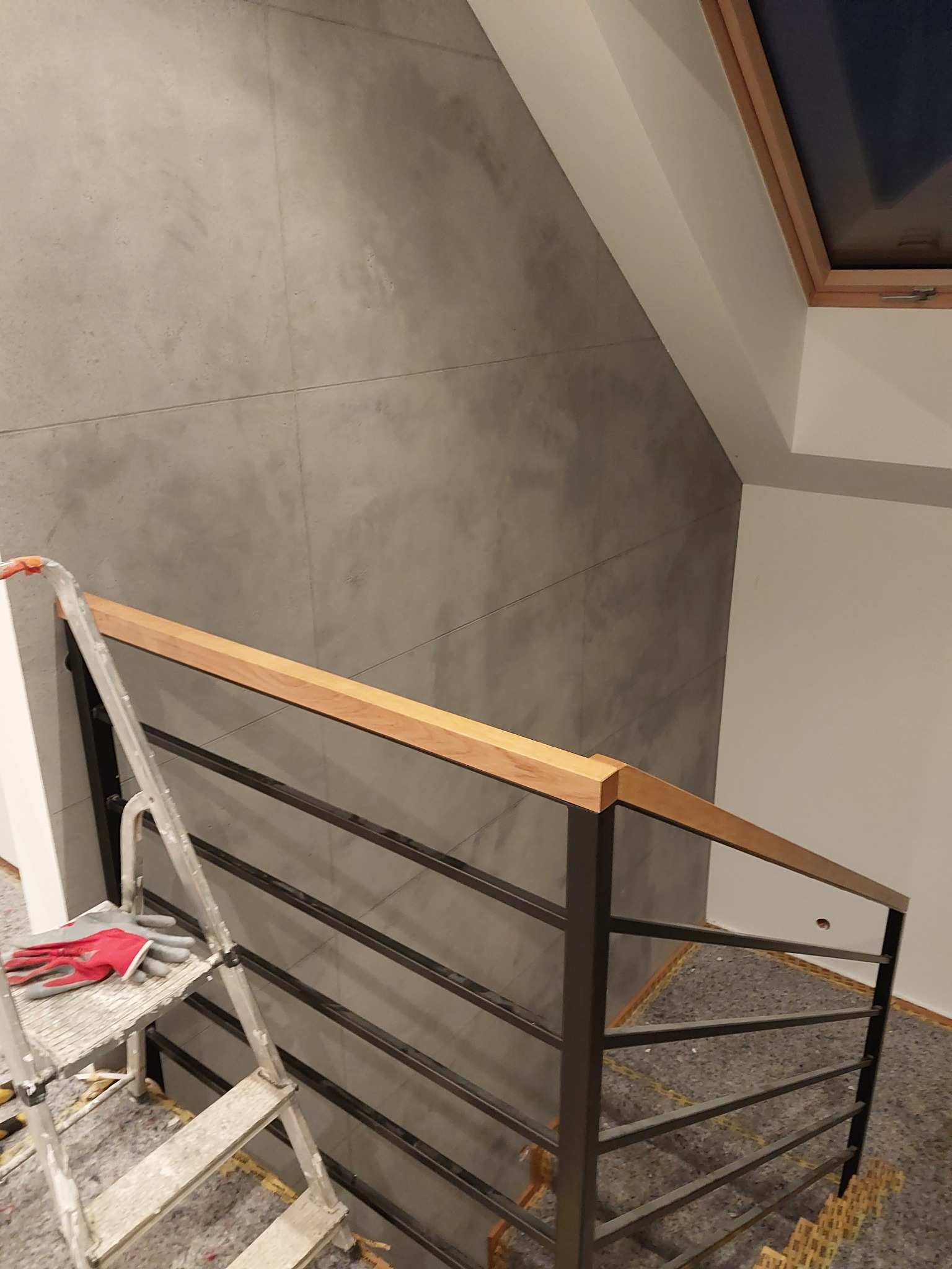 Ściana pokryta szarą, dekoracyjną fakturą przypominającą beton, widoczna z perspektywy schodów z metalową balustradą i drewnianym poręczą, drabina malarska z rękawicami na górnym stopniu, podłoga...