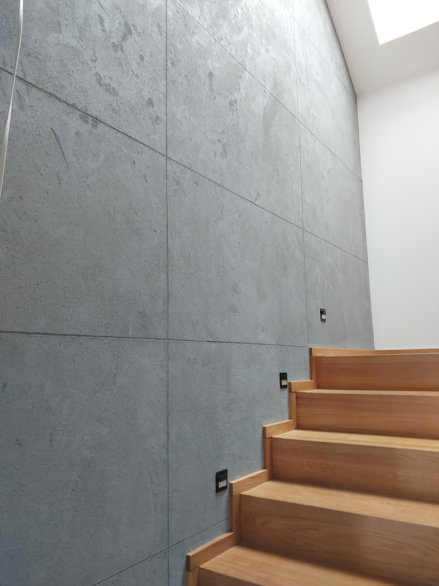 Ściana z szarego betonu architektonicznego ułożonego w kwadraty, oświetlenie schodowe przy drewnianych schodach, naturalne światło z góry.