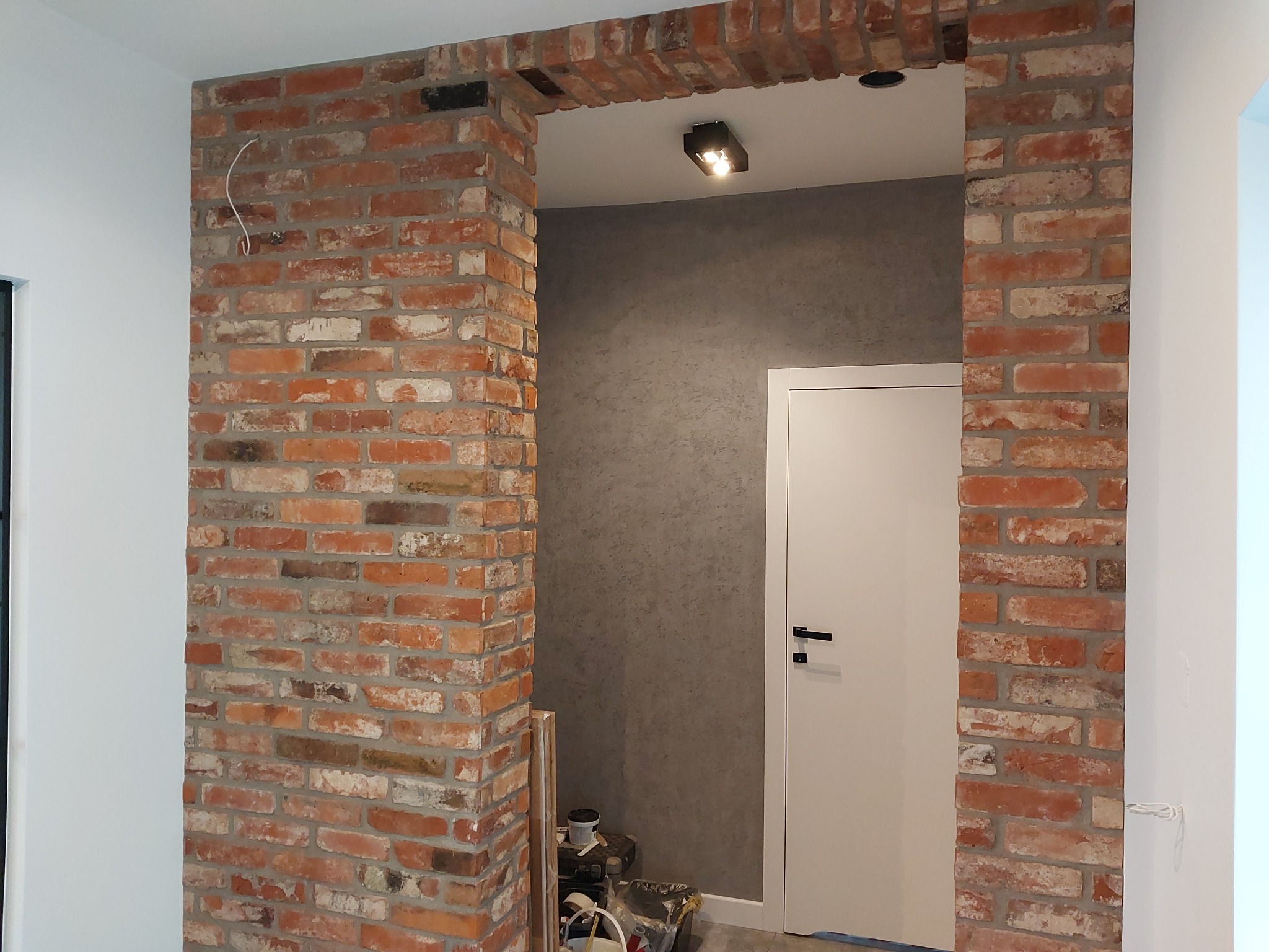 Aranżacja wnętrza z ceglanym łukiem nad drzwiami, surowa cegła kontrastuje z gładką, szarą ścianą i białą stolarką drzwiową, widoczne narzędzia i materiały budowlane sugerują etap wykończeniowy.
