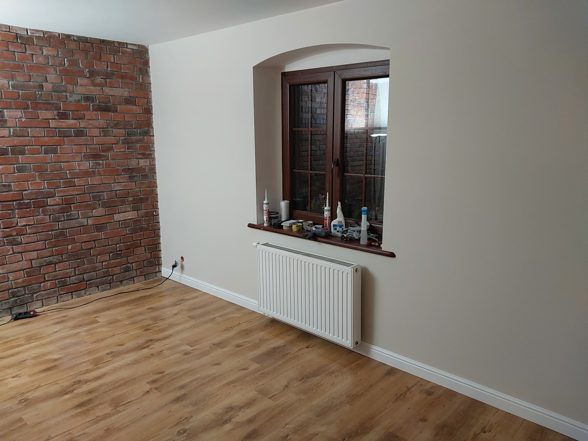 Pokój po remoncie z drewnopodobną podłogą, ścianą z cegły dekoracyjnej i oknem z brązową ramą. Na parapecie okiennym widoczne narzędzia i materiały budowlane.