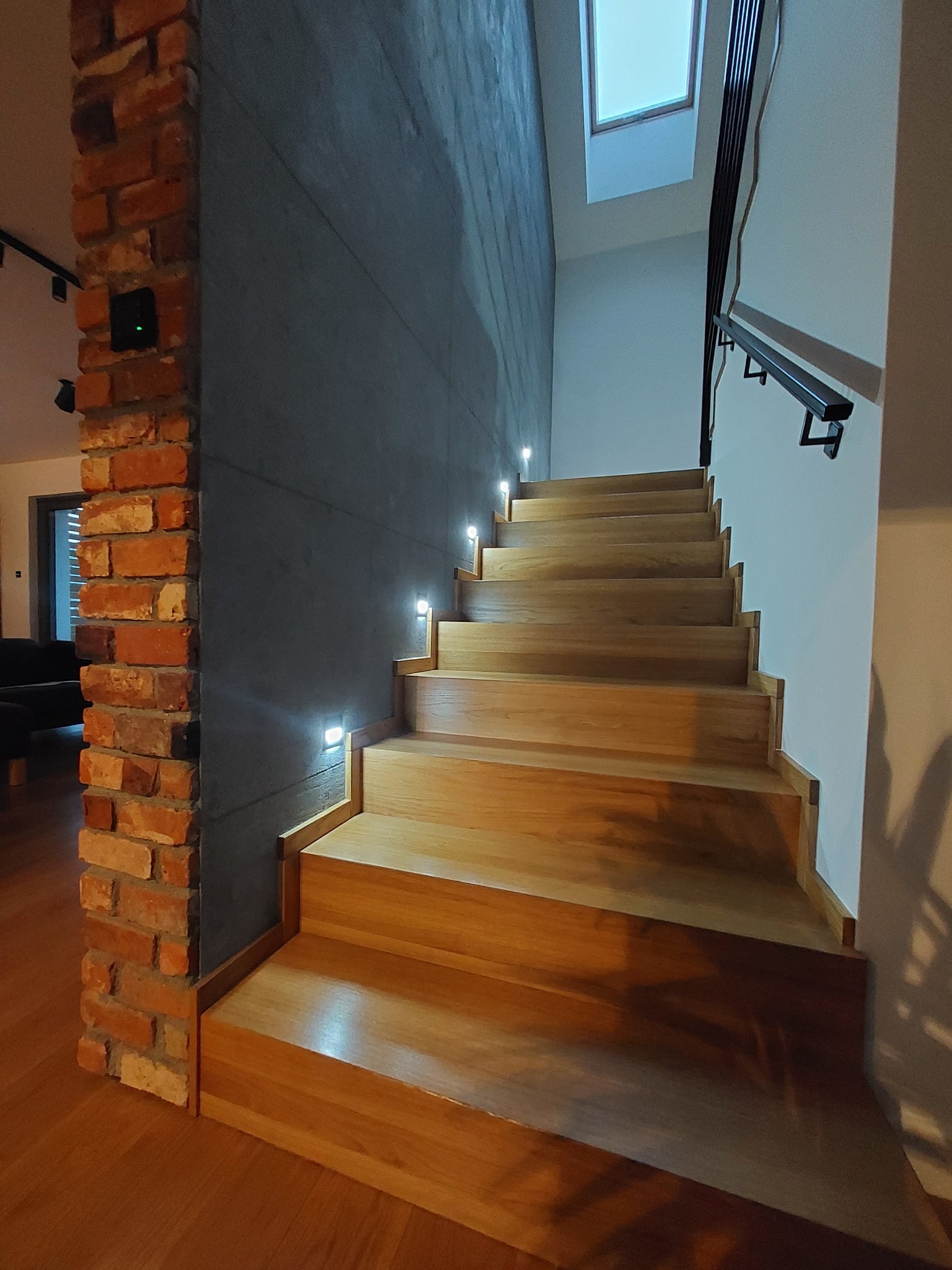 Nowoczesne schody drewniane z oświetleniem punktowym w ścianie o surowym wykończeniu, kolumna z cegły rozbiórkowej po lewej stronie, widok z dołu.