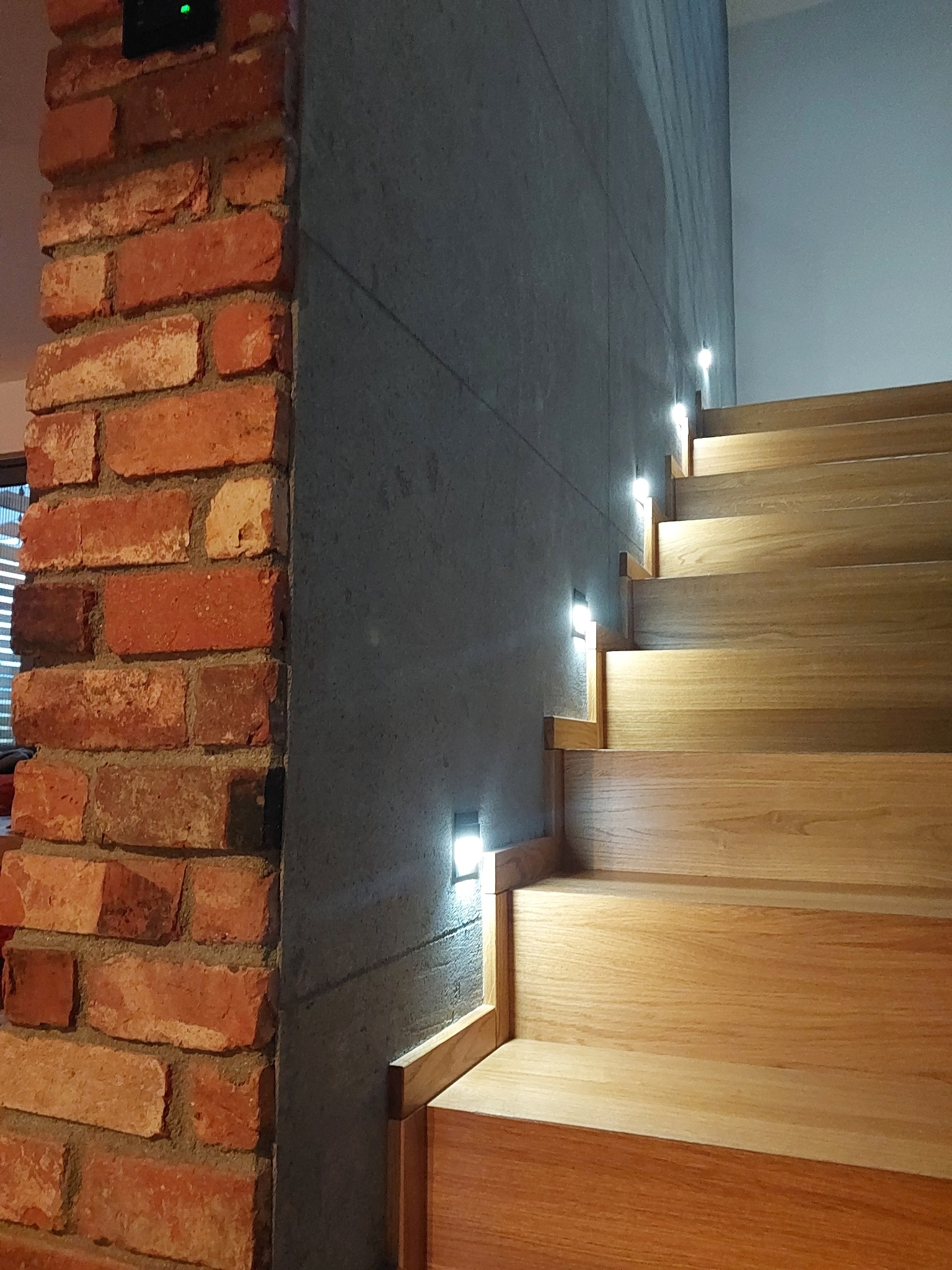 Nowoczesne schody drewniane z oświetleniem LED wzdłuż betonowej ściany i dekoracyjnej ściany z cegły, Siedlisko.