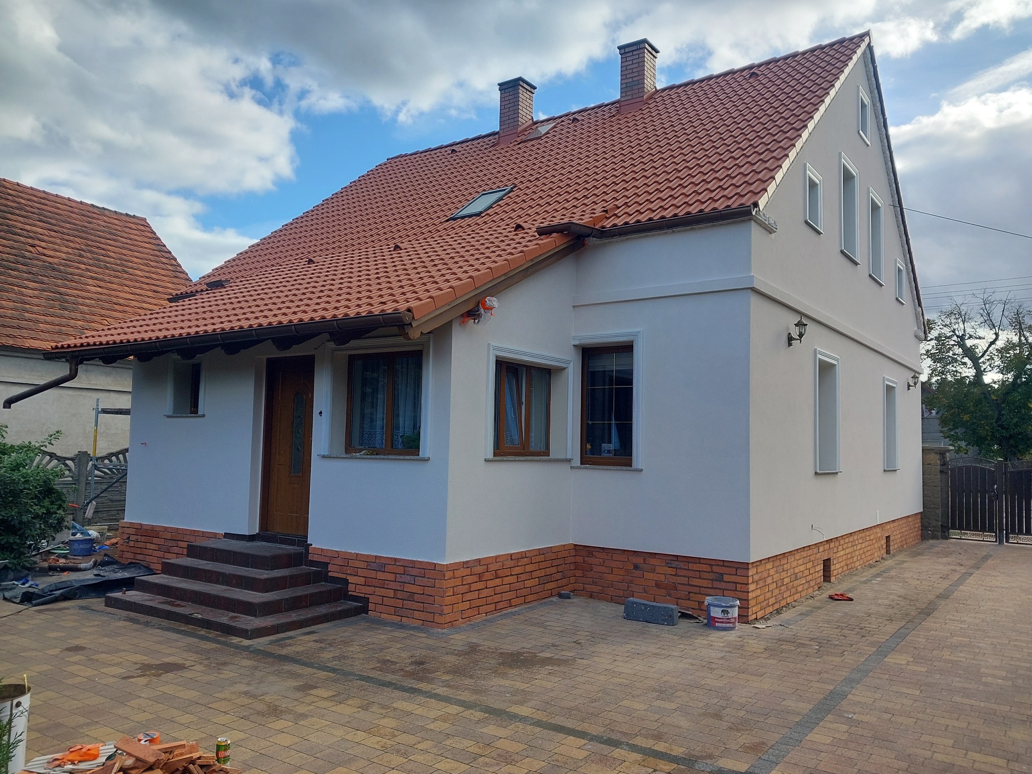 Odnowiony dom jednorodzinny z nową elewacją w kolorze jasnego beżu, czerwoną dachówką i ceglanym cokołem, widok z chodnika z kostki brukowej w słoneczny dzień.