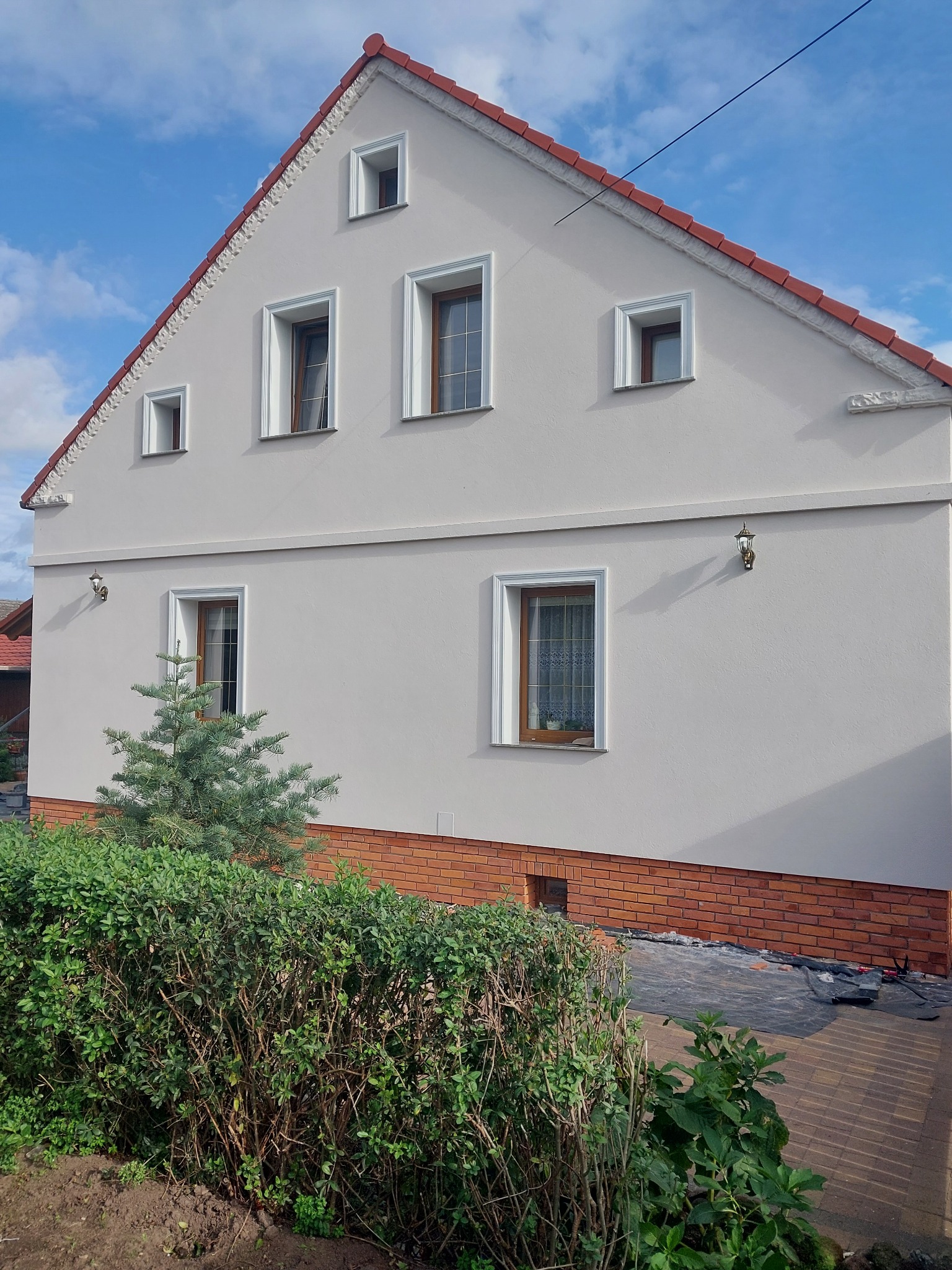 Wykończona elewacja domu z jasnoszarym tynkiem, dekoracyjnymi elementami pod dachem i ceglaną podmurówką, widok z poziomu ogrodu z krzewami i małą choinką.