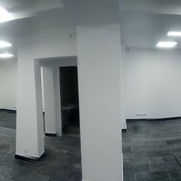 Usługi Elektryczne Paweł Małocha - Panoramiczne ujęcie wnętrza po montażu oświetlenia sufitowego, z widocznymi grzejnikami pod oknami i ciemną podłogą z płytek, w trakcie prac wykończeniowych.
