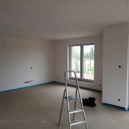 Usługi Elektryczne Paweł Małocha - Wnętrze w stanie deweloperskim z widocznymi punktami elektrycznymi na ścianie, przygotowanymi do montażu oświetlenia. Na podłodze stoi aluminiowa drabina, a kable zwisają z sufitu.