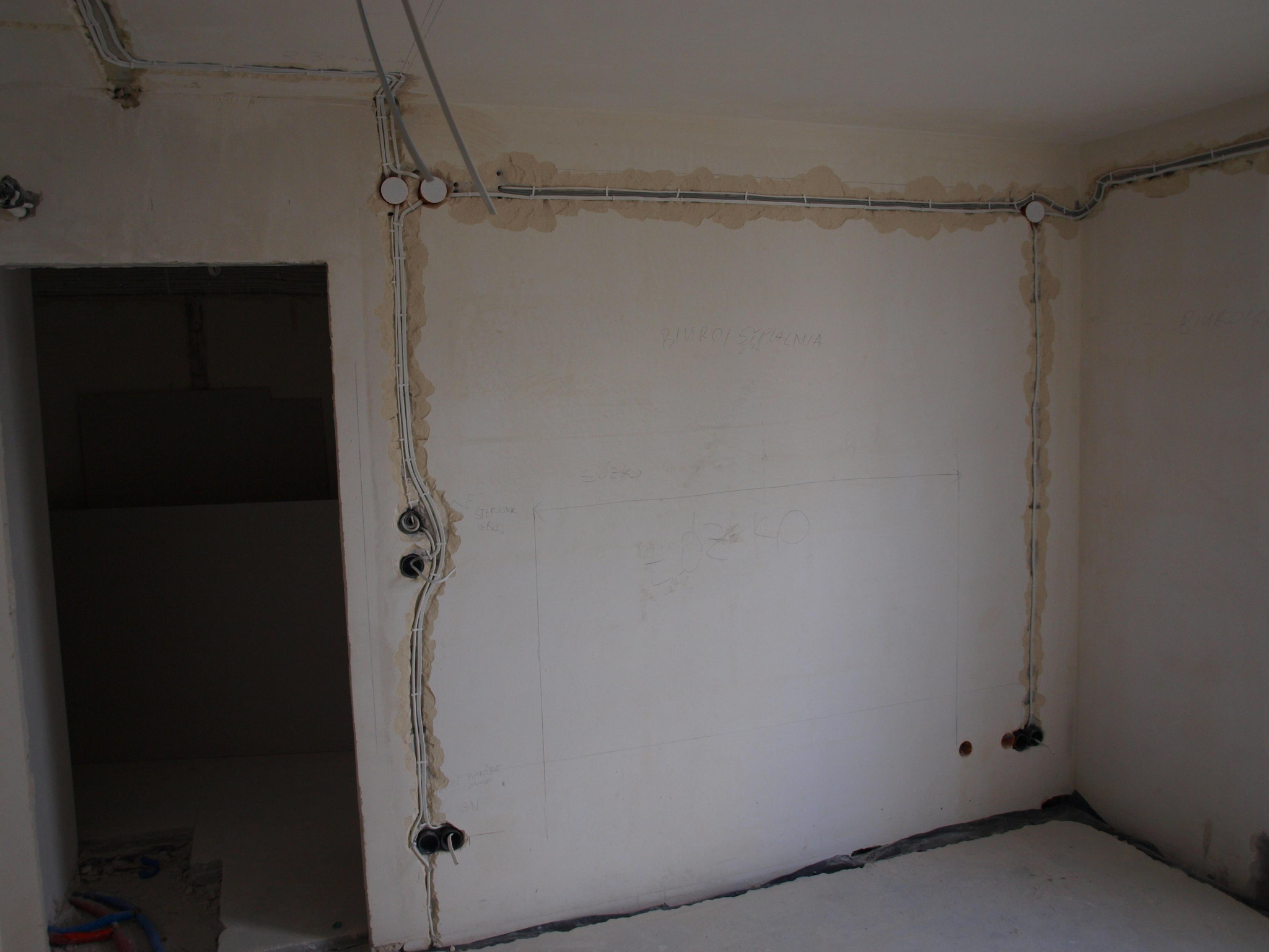 Surowy stan pokoju z widoczną instalacją elektryczną w ścianach, obejmującą kable w peszlach i puszki podtynkowe, przygotowaną do dalszych prac wykończeniowych.