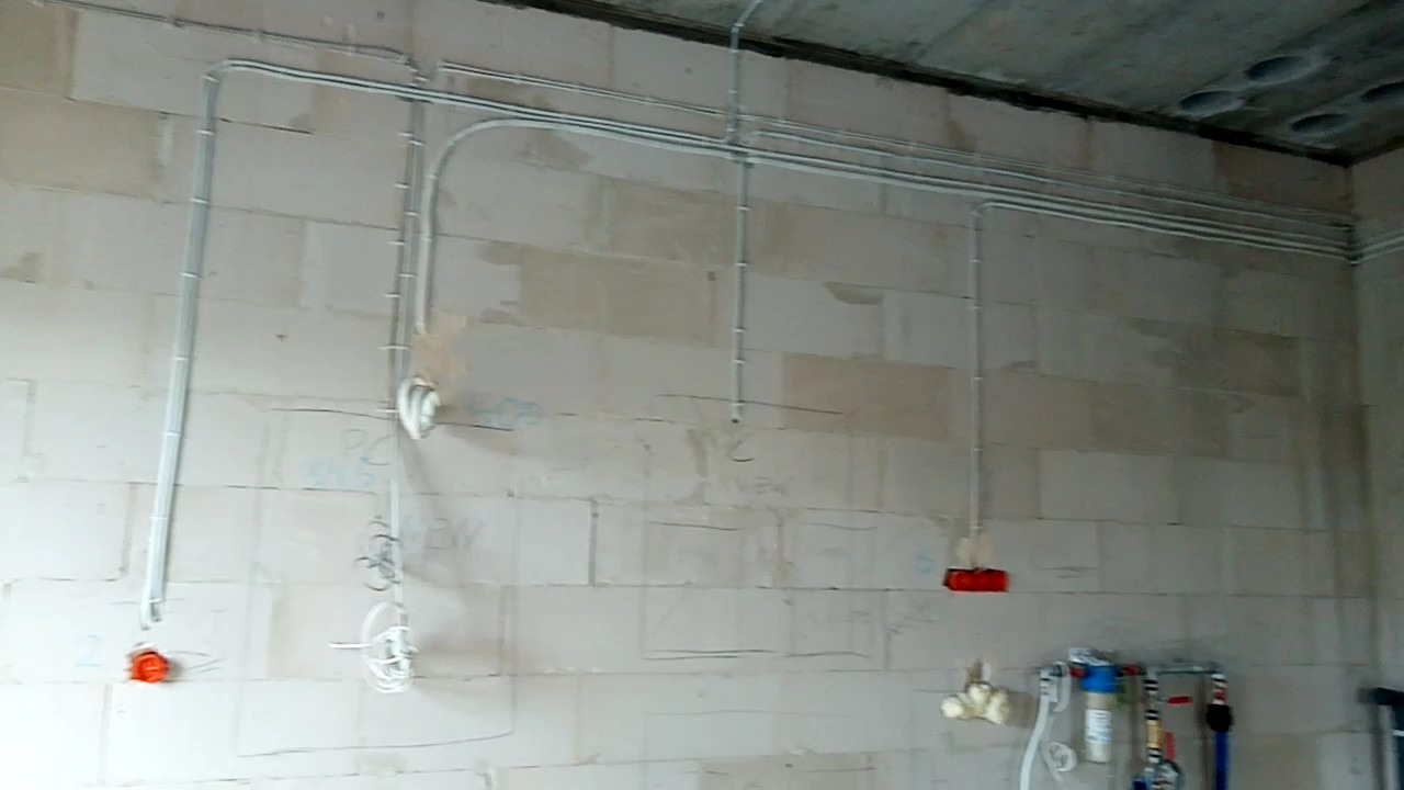 Szare rury elektryczne prowadzone po ścianie z białych bloczków w surowym stanie, widoczne puszki instalacyjne w kolorze pomarańczowym i białe kable zwisające ze ściany, instalacja wodna w dolnym...