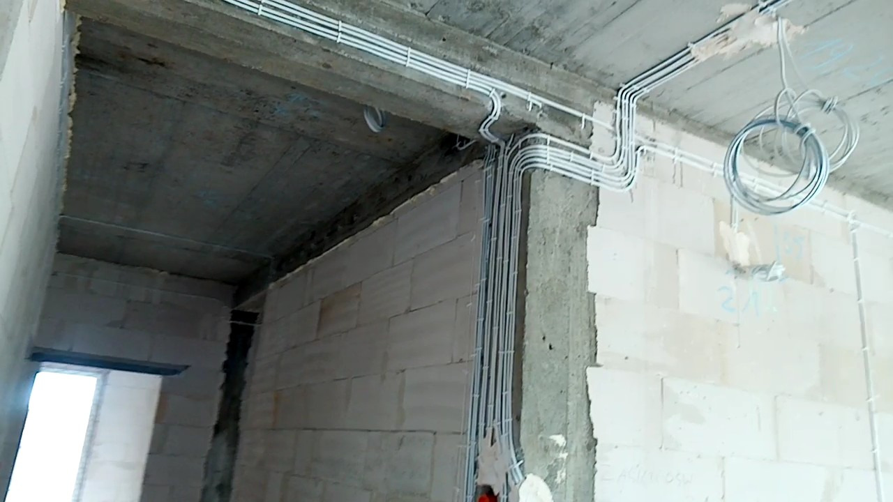 Surowy stan budynku w trakcie prac: białe rurki instalacji elektrycznej prowadzone po suficie i ścianach z białego bloczka, zwisający kabel, widoczne napisy na ścianie.