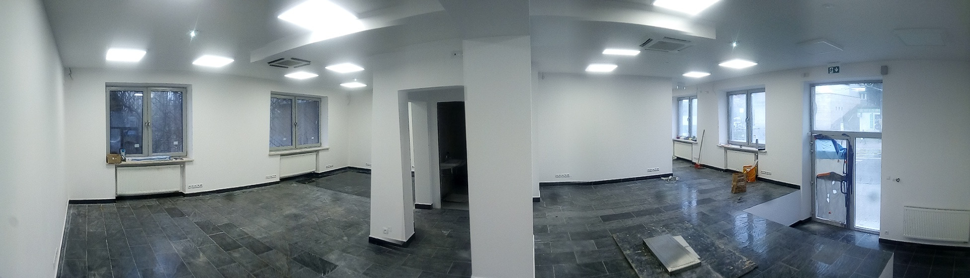 Panoramiczne ujęcie wnętrza po montażu oświetlenia sufitowego, z widocznymi grzejnikami pod oknami i ciemną podłogą z płytek, w trakcie prac wykończeniowych.