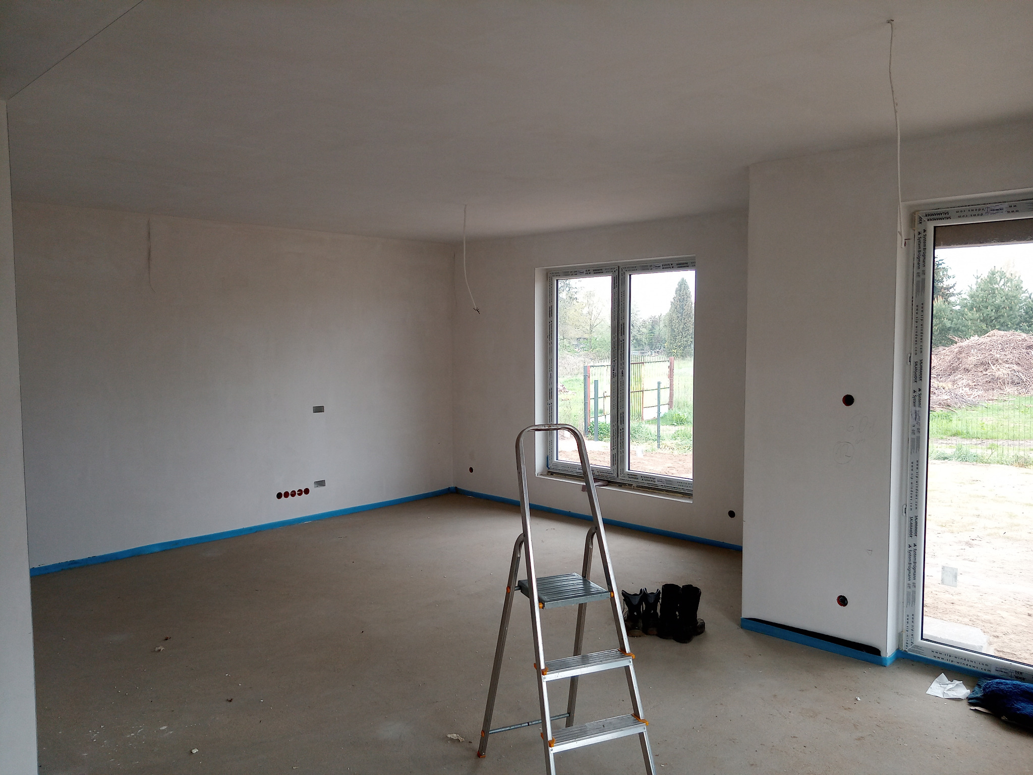 Wnętrze w stanie deweloperskim z widocznymi punktami elektrycznymi na ścianie, przygotowanymi do montażu oświetlenia. Na podłodze stoi aluminiowa drabina, a kable zwisają z sufitu.