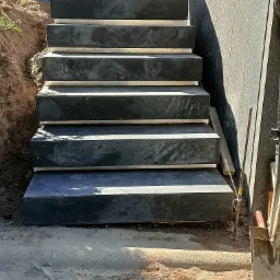 Betonowe schody zewnętrzne w trakcie budowy, widoczne narzędzia: łopata, poziomica, ziemia wokół.