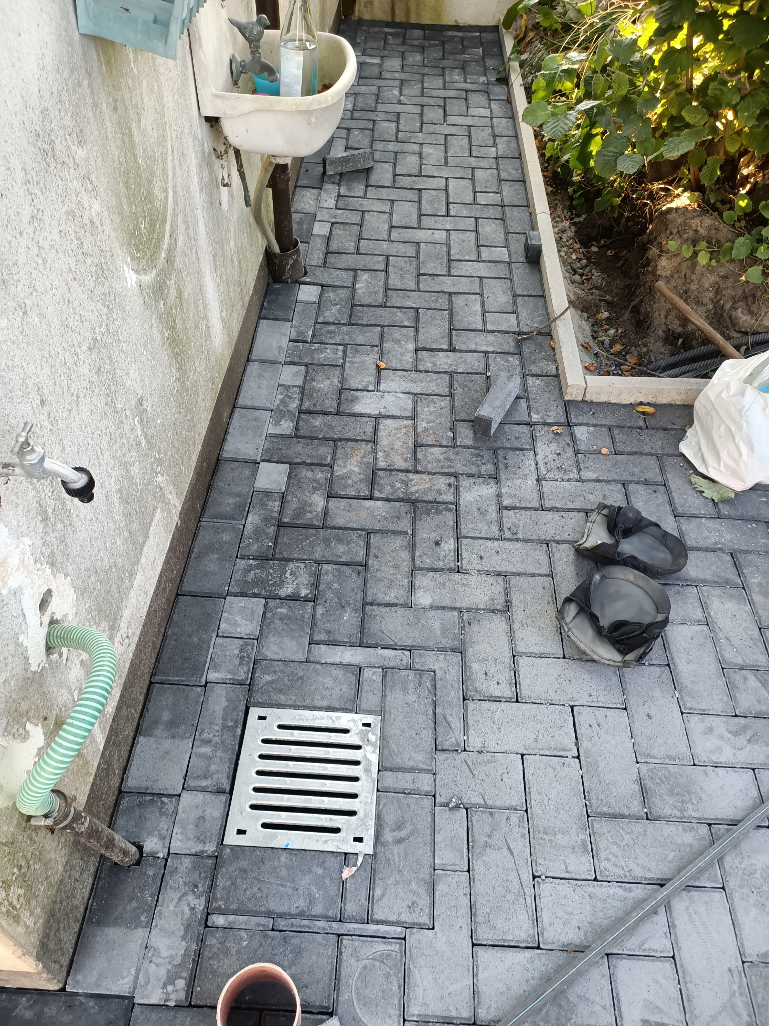 Świeżo ułożona, szara nawierzchnia z kostki, z widocznym odpływem liniowym, kolanami ochronnymi i fragmentem ściany z kranem.