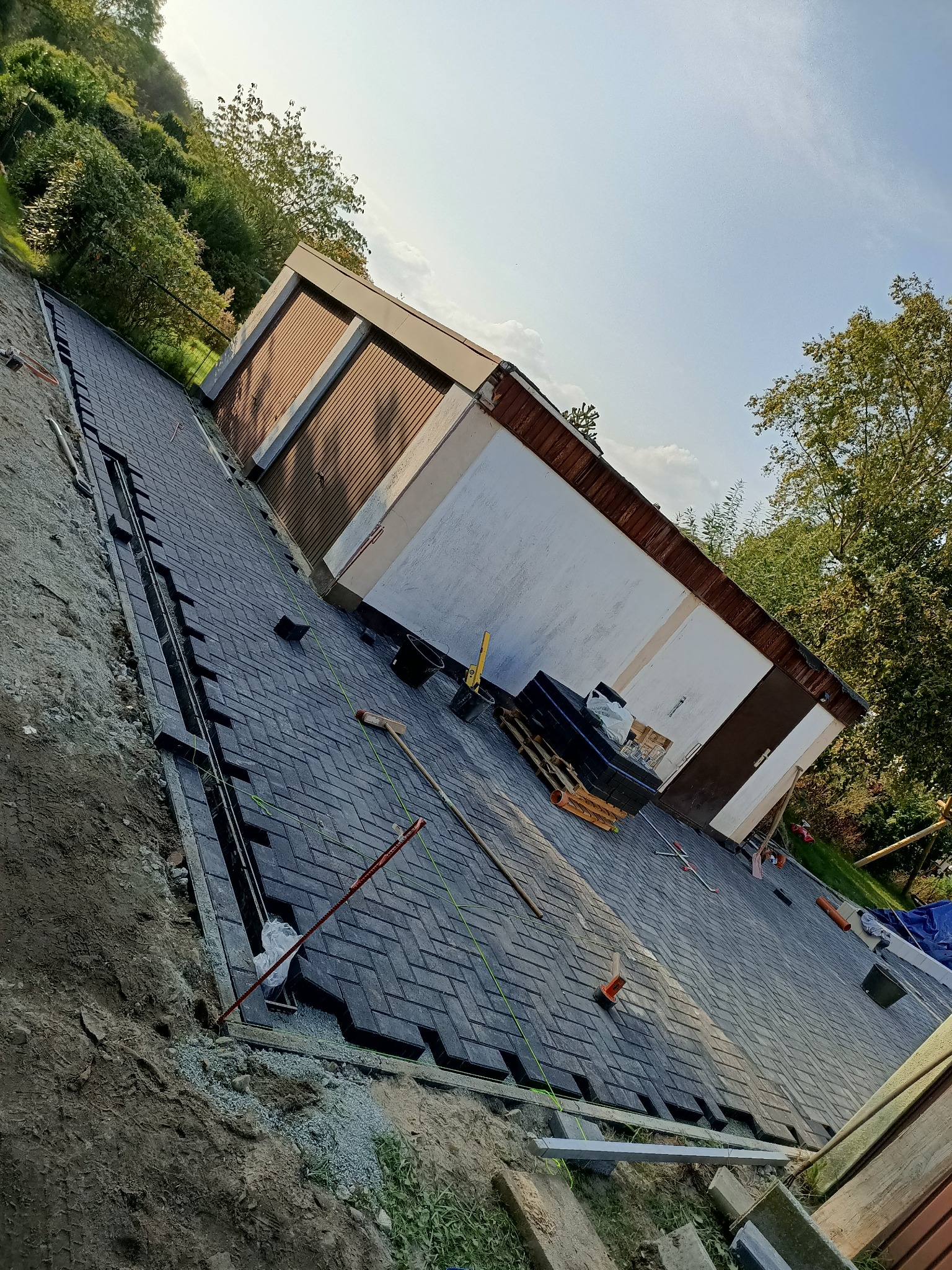 Ciemnoszara kostka ułożona we wzór jodełki na podjeździe, z widocznym garażem w tle i narzędziami pracy na powierzchni.