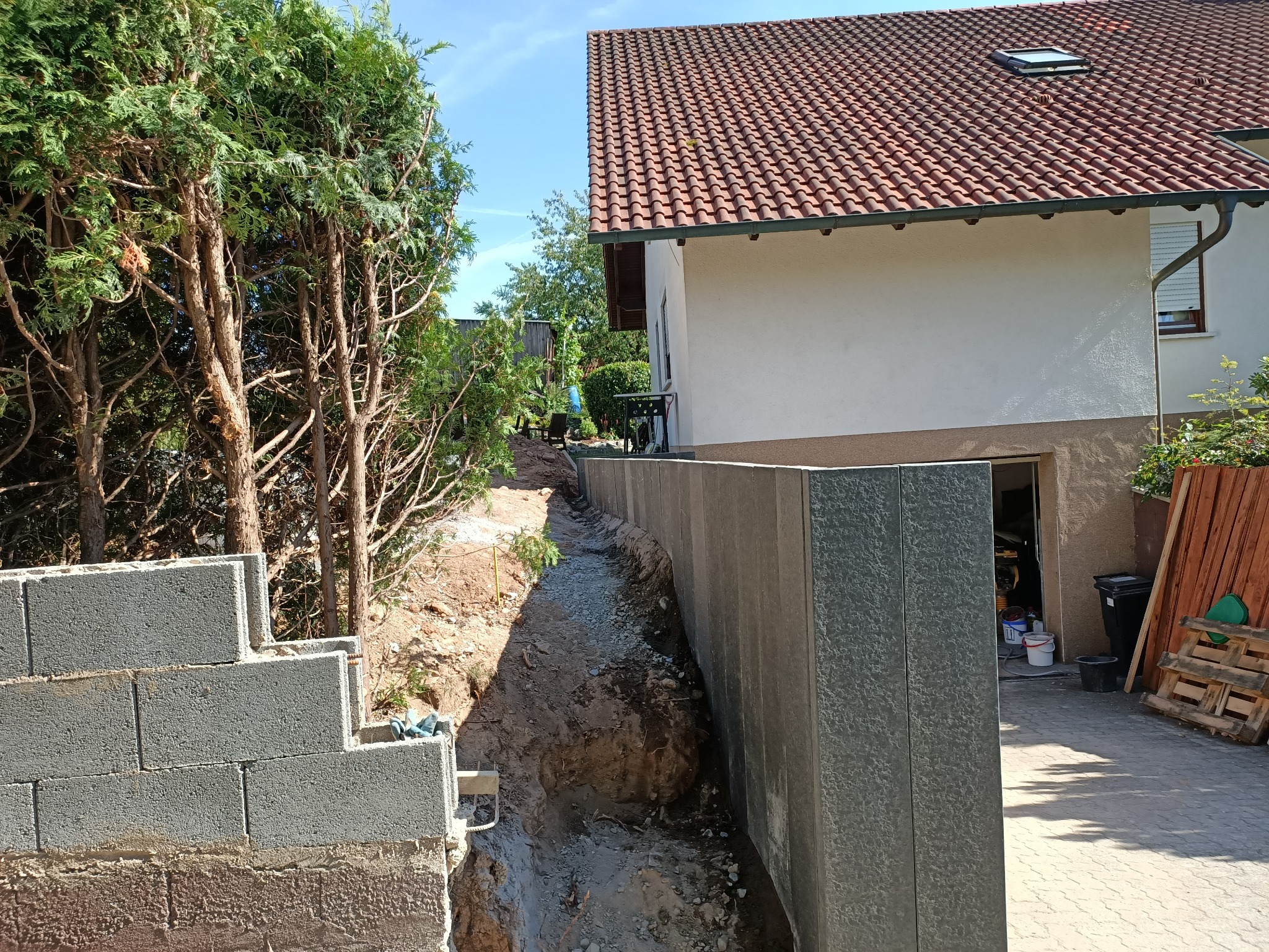 Budowa muru oporowego z palisady betonowej na nierównym terenie, widoczne bloczki betonowe i wykop pod fundament, w tle dom z czerwoną dachówką.