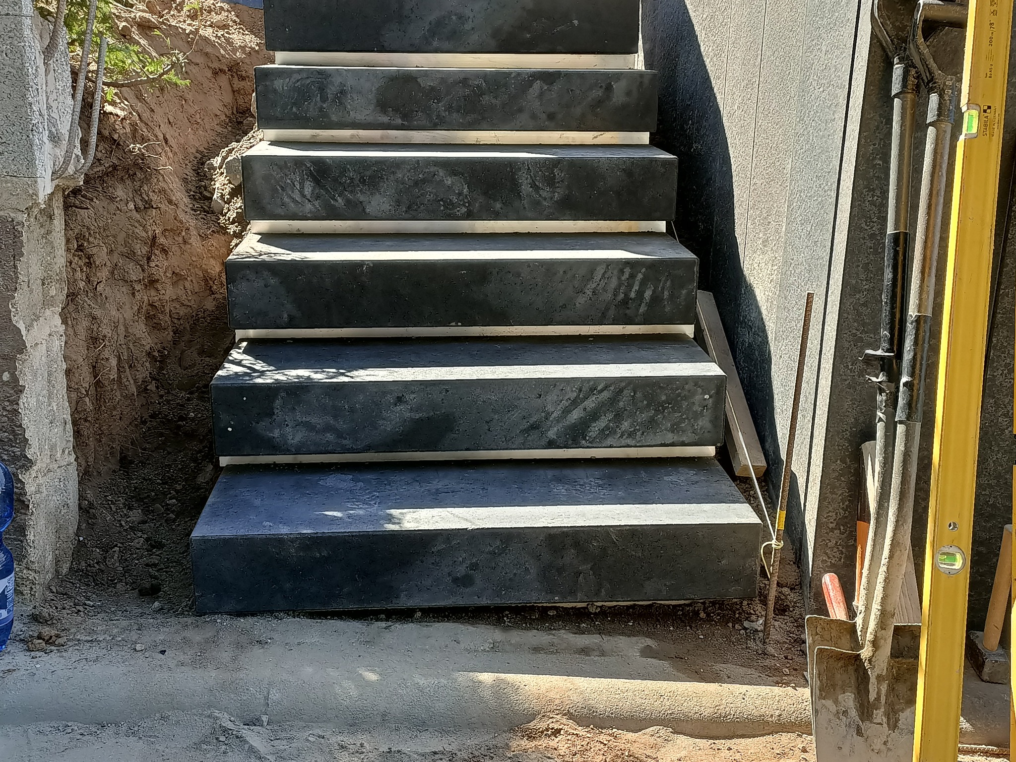 Betonowe schody zewnętrzne w trakcie budowy, widoczne narzędzia: łopata, poziomica, ziemia wokół.