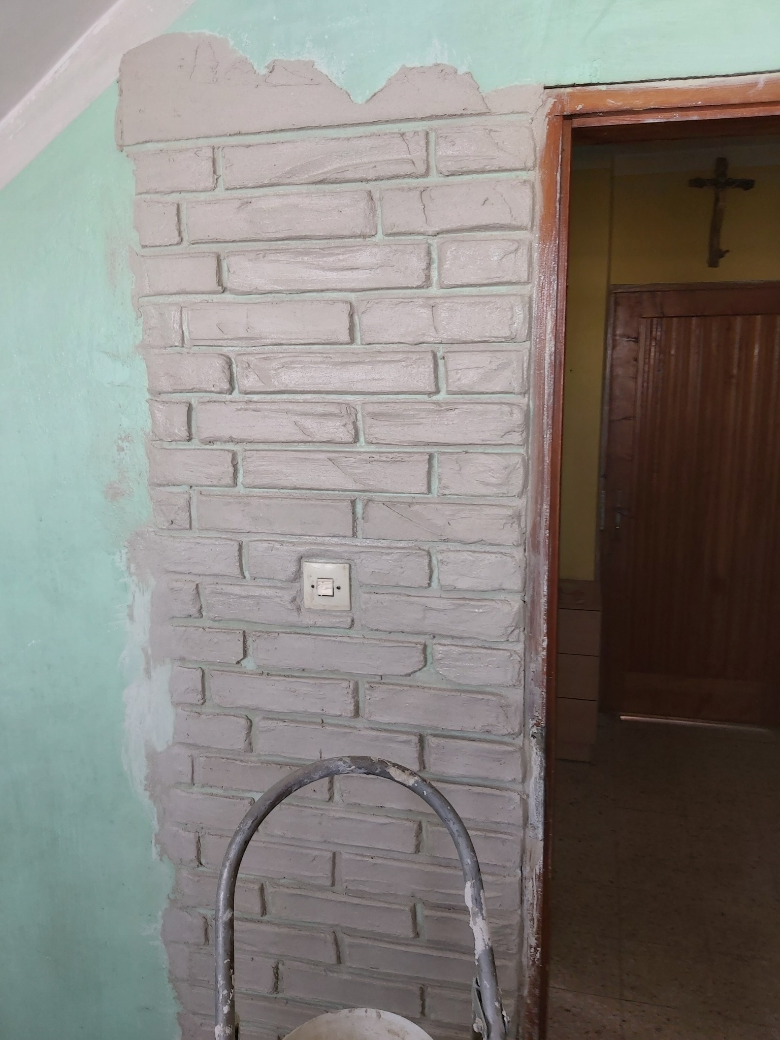 Ściana z imitacją cegły w trakcie wykańczania, widoczny fragment pomalowanej ściany na zielono i fragment nieukończonej imitacji cegły obok futryny drzwi.