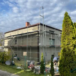 Dwukondygnacyjny budynek w trakcie prac elewacyjnych, otoczony rusztowaniem, z widocznym pracownikiem przy ziemi i gęstymi tujami w pierwszym planie.