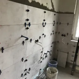 Ściana w trakcie układania płytek ceramicznych z użyciem systemu poziomowania, widoczne otwory na instalacje oraz poziomica przy oknie.