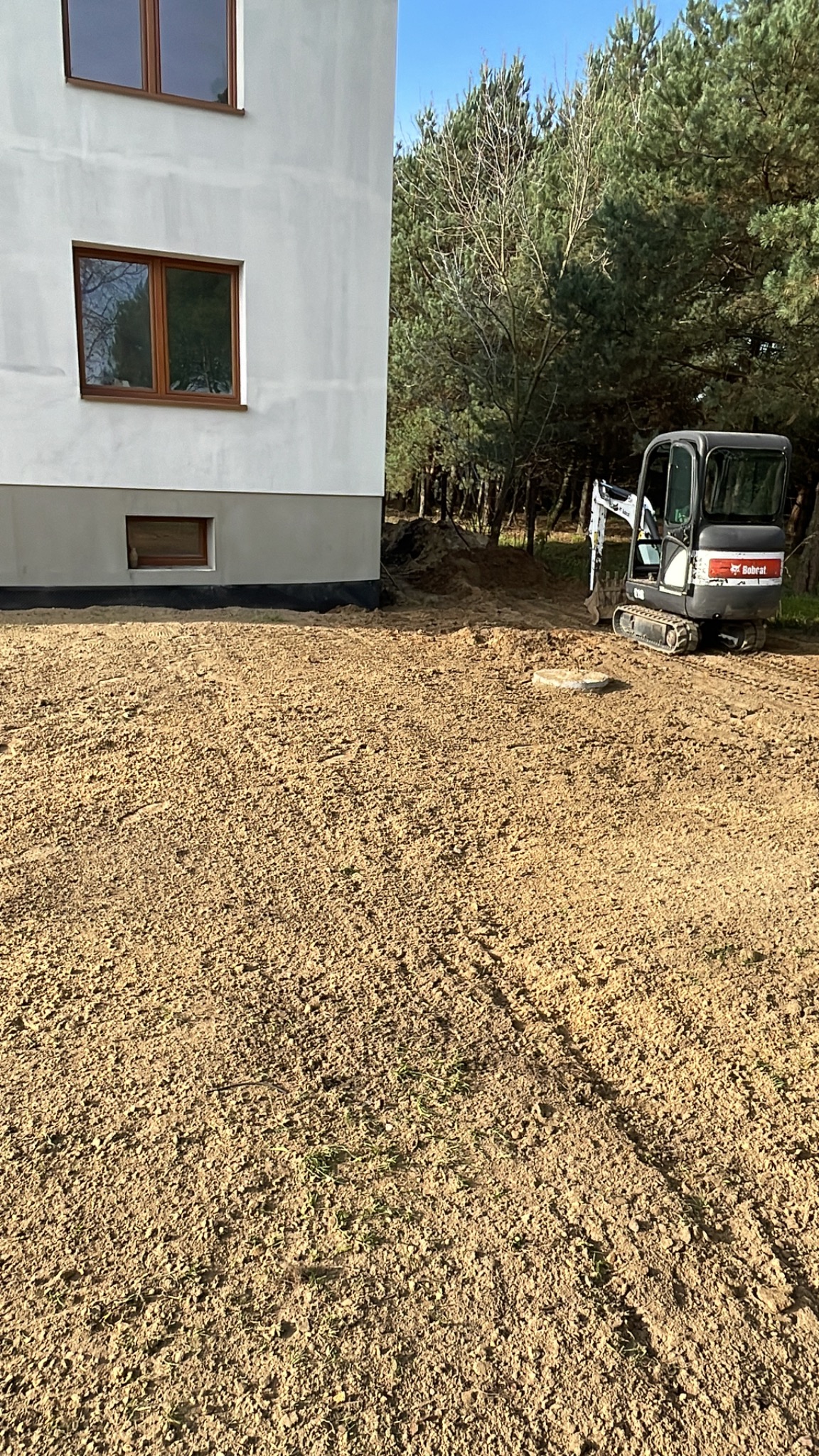 Przygotowany teren pod budowę domu jednorodzinnego z widocznym mini-koparką Bobcat i wystającą studzienką kanalizacyjną.