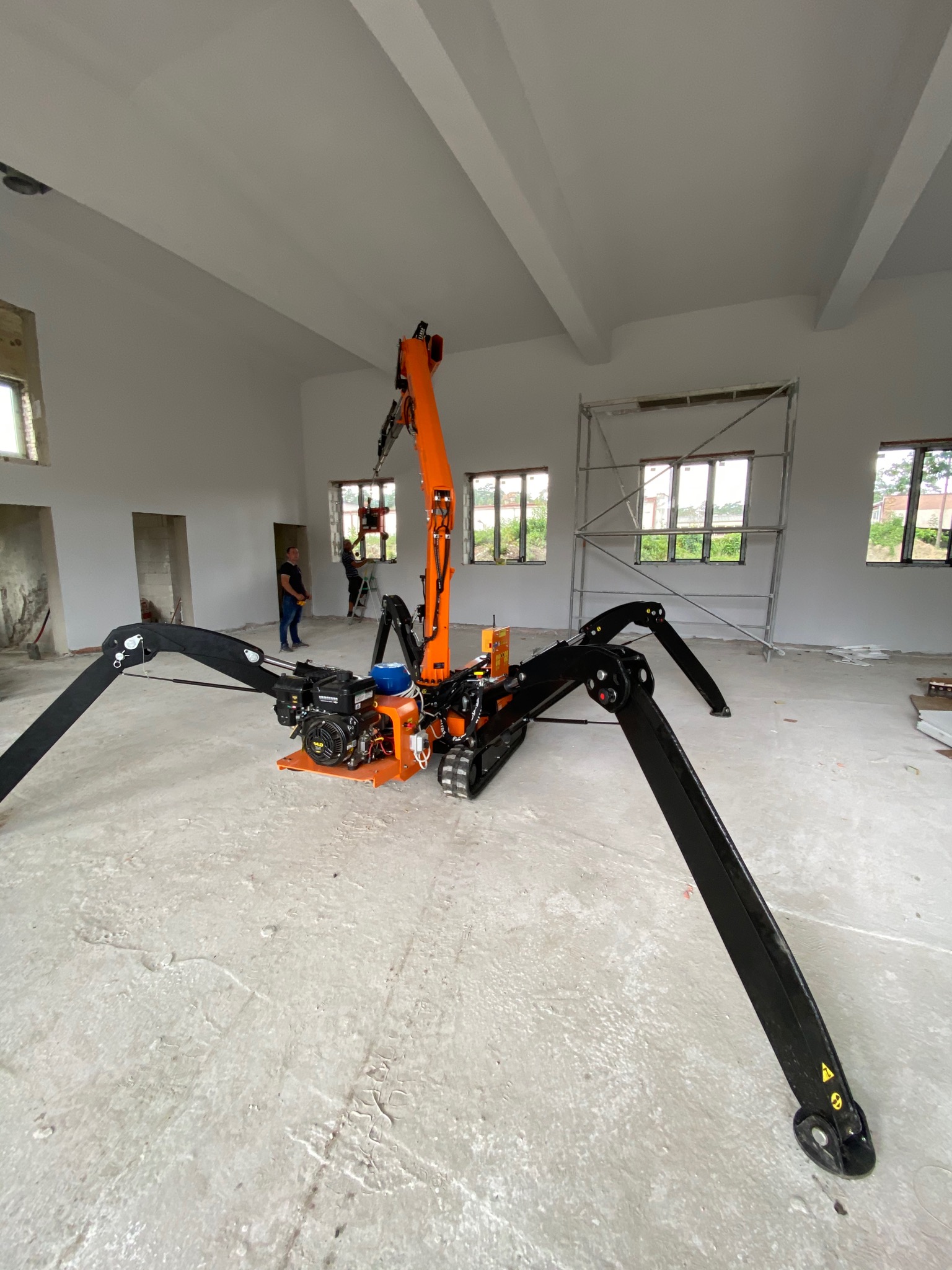 Pomarańczowo-czarny pająk-dźwig na gąsienicach z rozłożonymi podporami w surowym wnętrzu, pracownicy montują okno na rusztowaniu w tle.